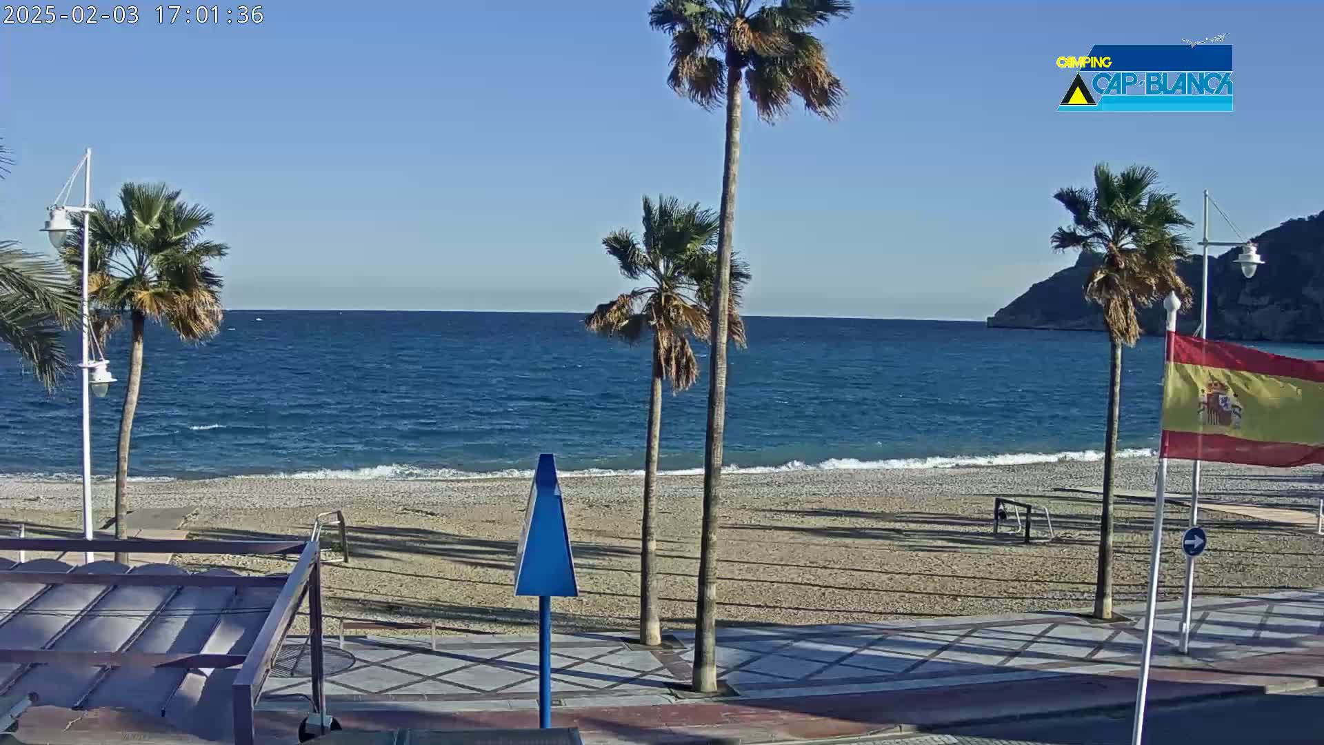A sunny beach scene shows a calm blue ocean lapping a pebble shore, lined with palm trees and a Spanish flag.