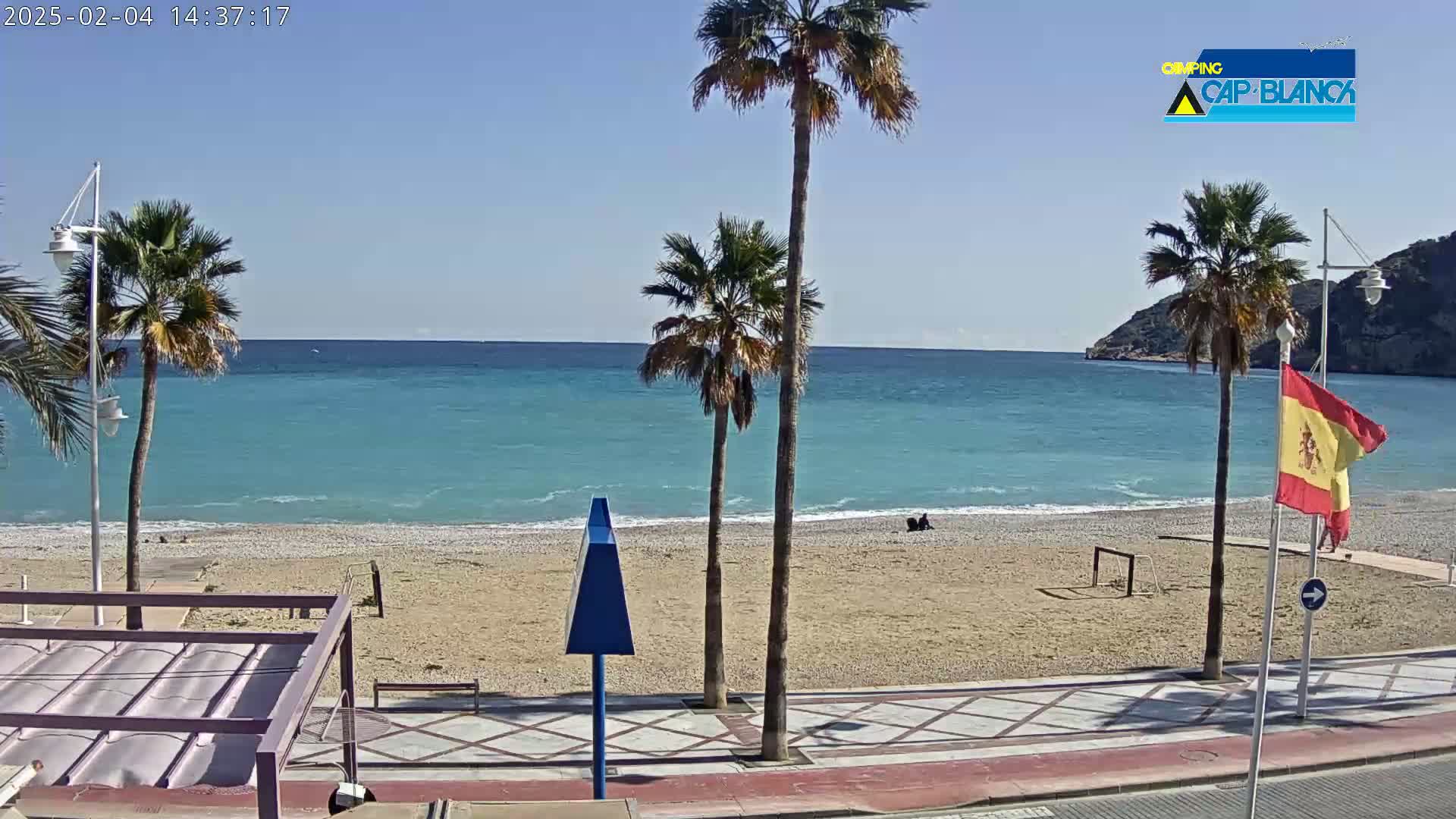 A sandy beach with palm trees and a calm, turquoise ocean under a clear blue sky is seen from a seaside promenade.