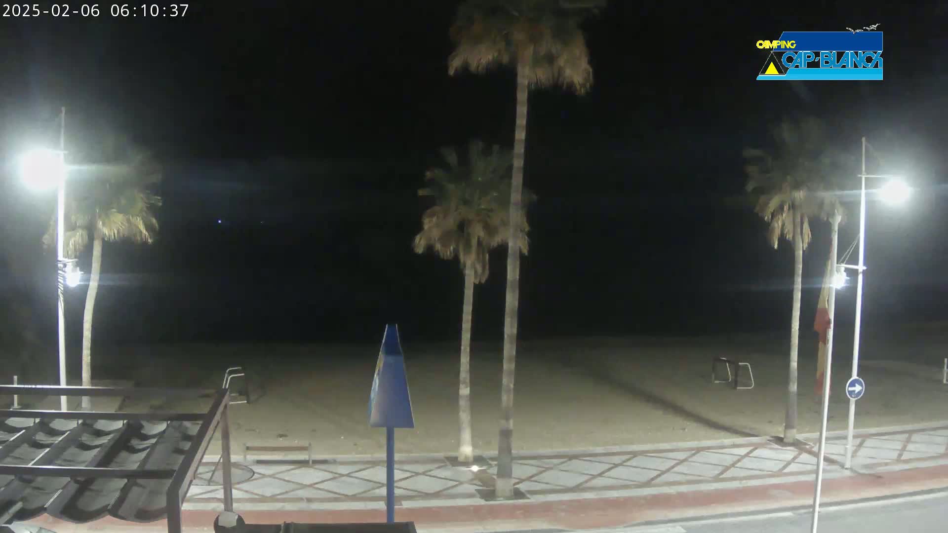 At night, a sandy beach is visible under bright lights, with palm trees lining a paved walkway.