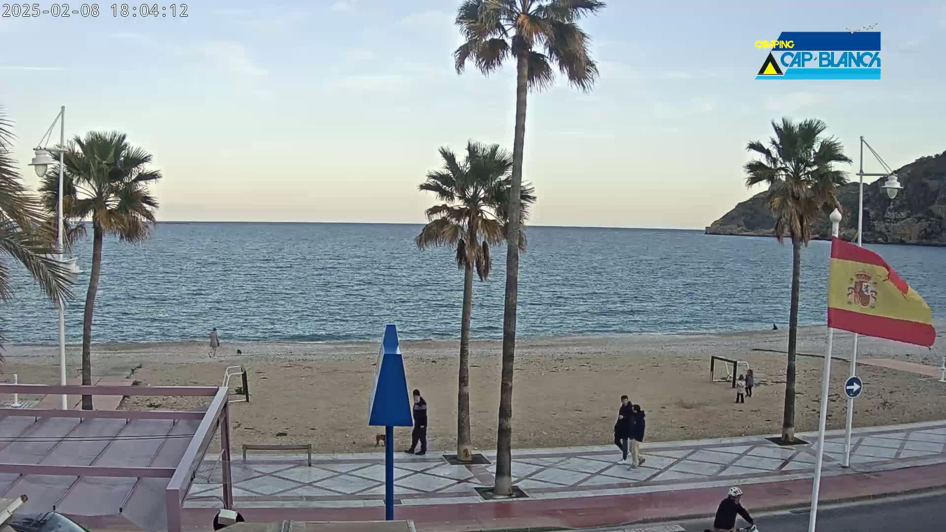 A mostly sunny beach scene shows palm trees, several people walking along a promenade next to a sandy beach and calm ocean, with a Spanish flag flying near a rocky headland.