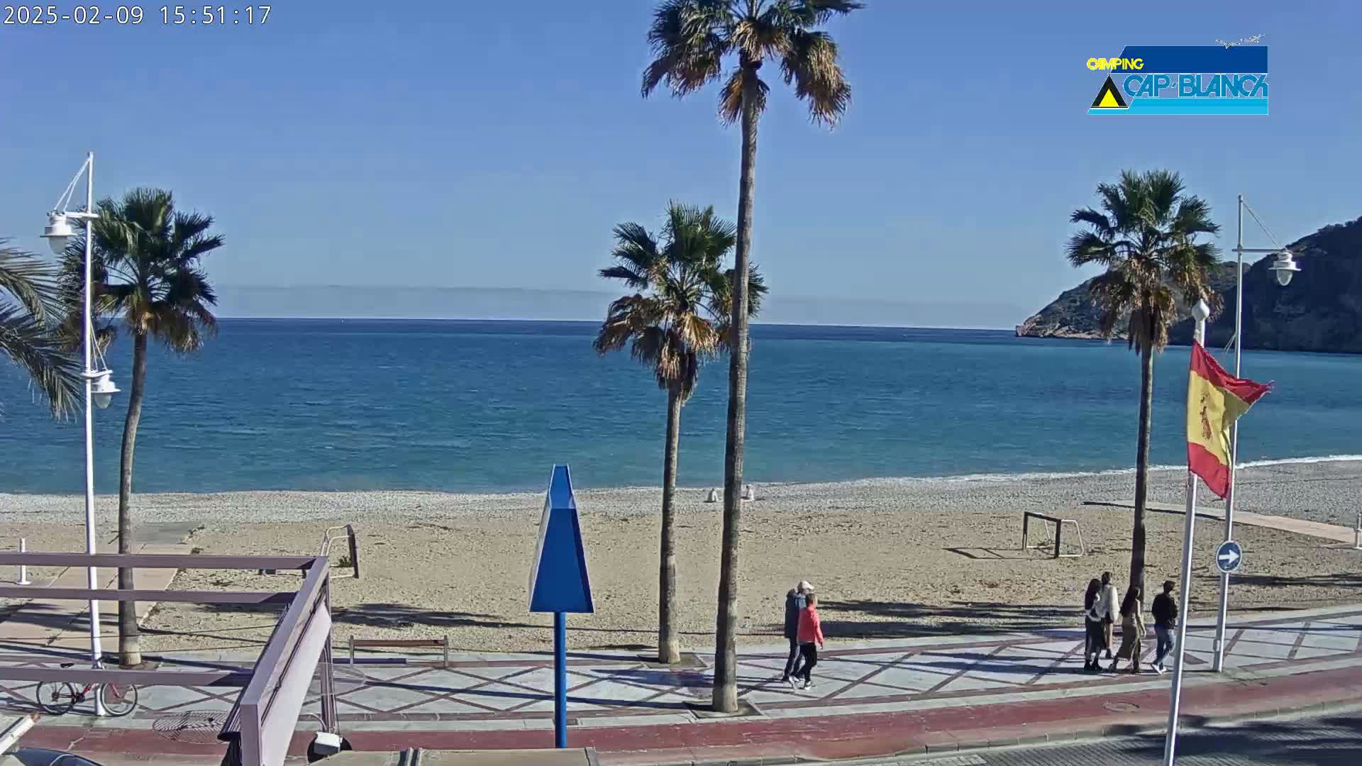 A sandy beach with palm trees and a few people walking along a promenade, overlooks a calm blue sea under a clear sky.