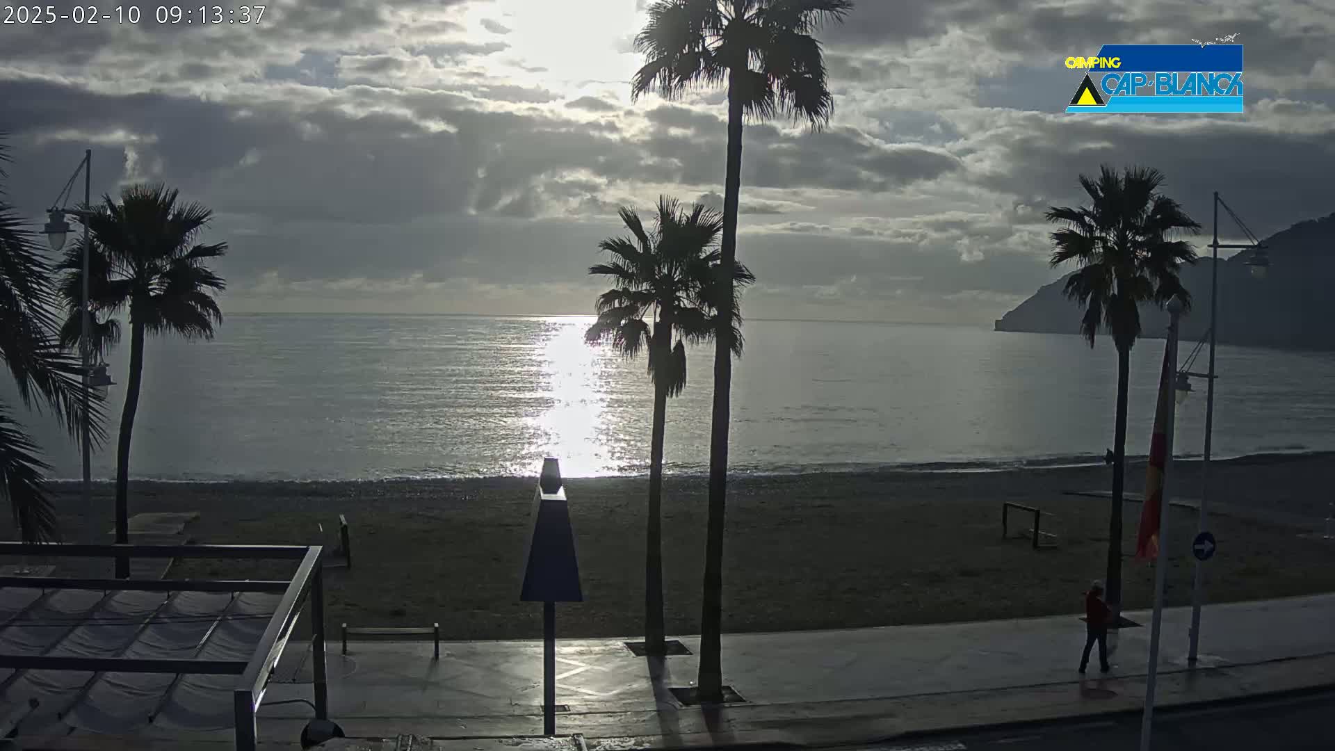 A mostly cloudy sunrise illuminates a calm sea with a sandy beach, palm trees, and a person walking along a promenade.