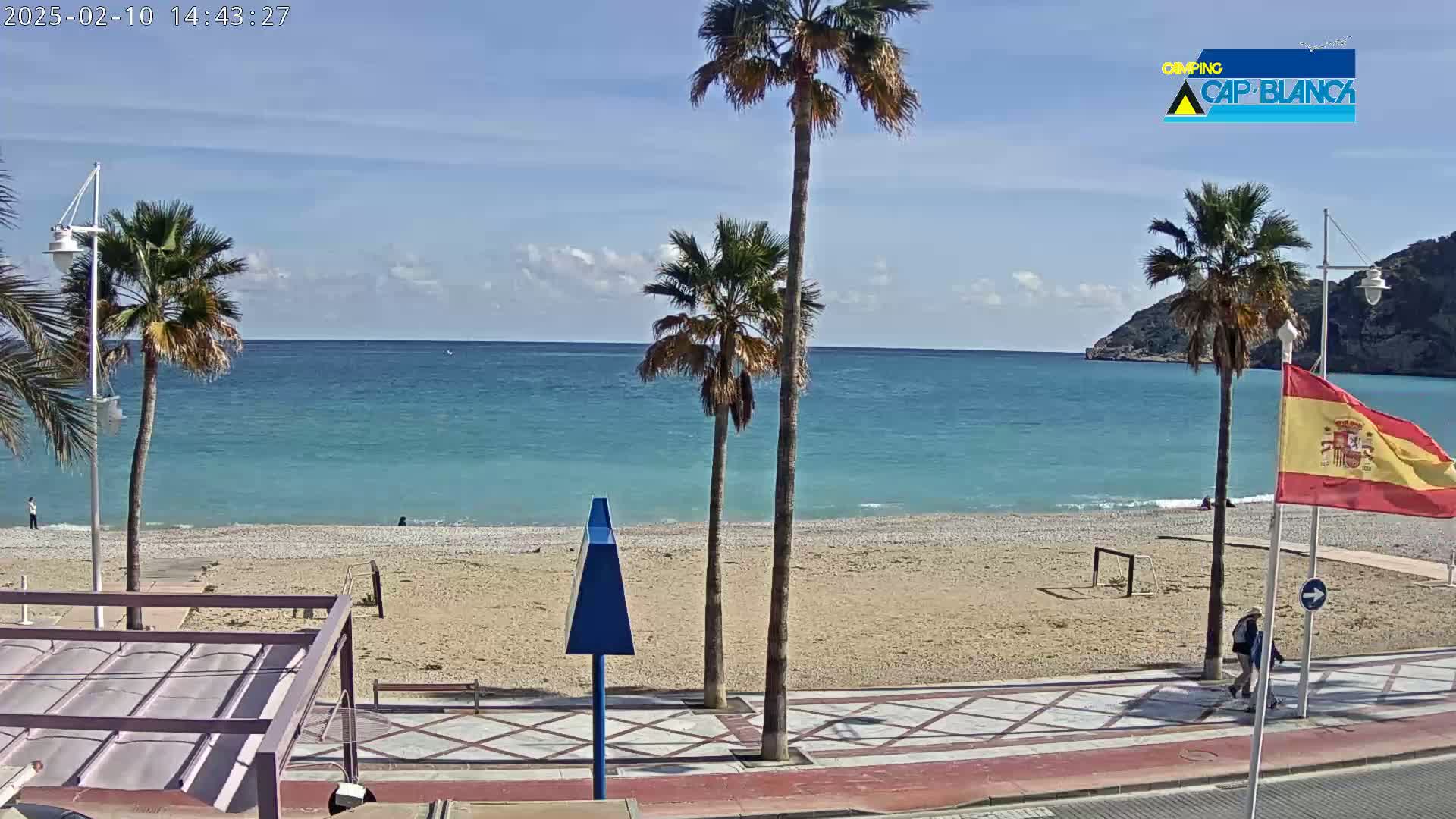A sandy beach with palm trees and a calm, turquoise ocean under a mostly sunny sky is viewed from a boardwalk.