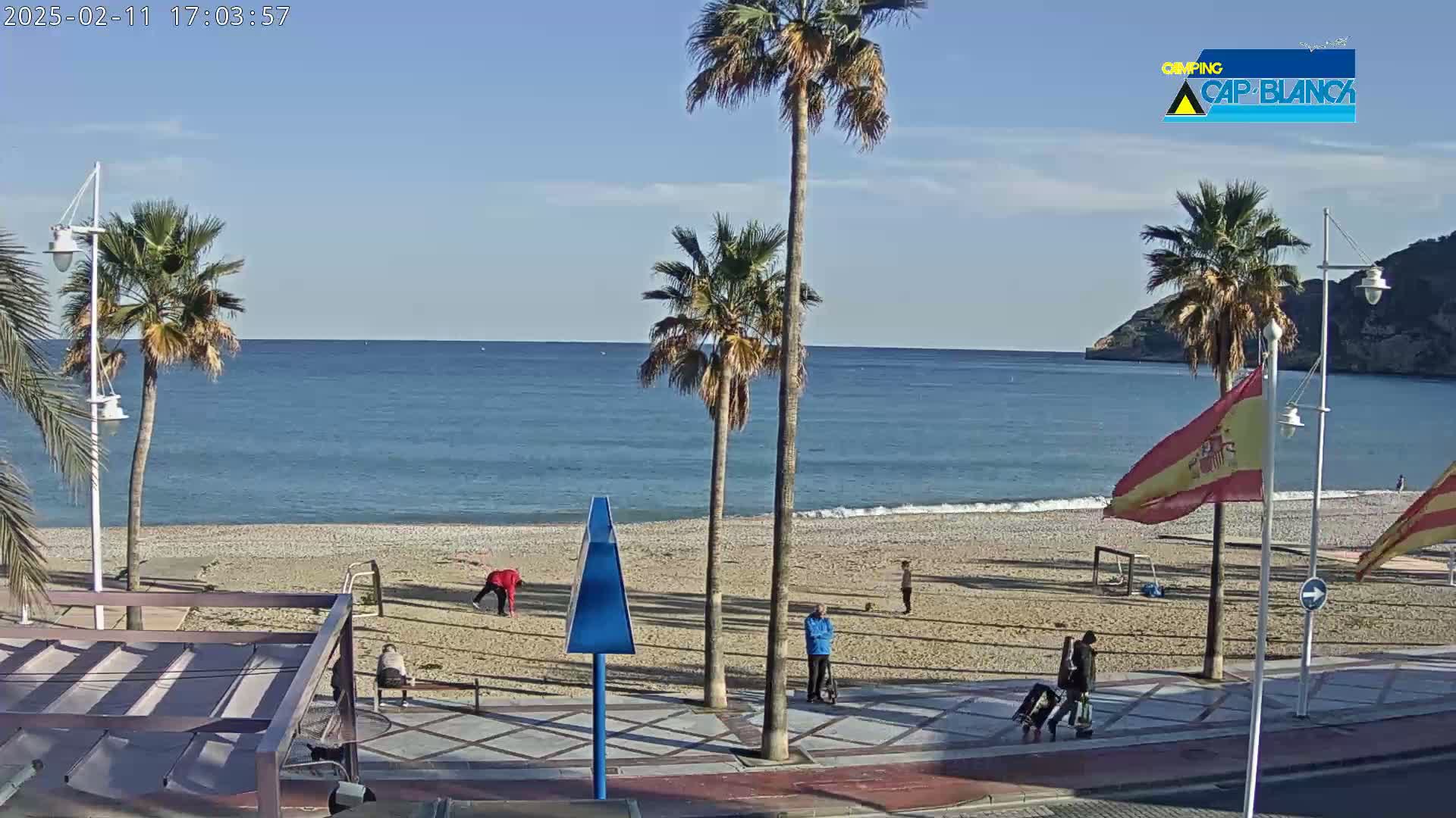A mostly sunny beach scene shows palm trees lining a sandy shore next to a calm blue sea, with several people walking along the beach and boardwalk.