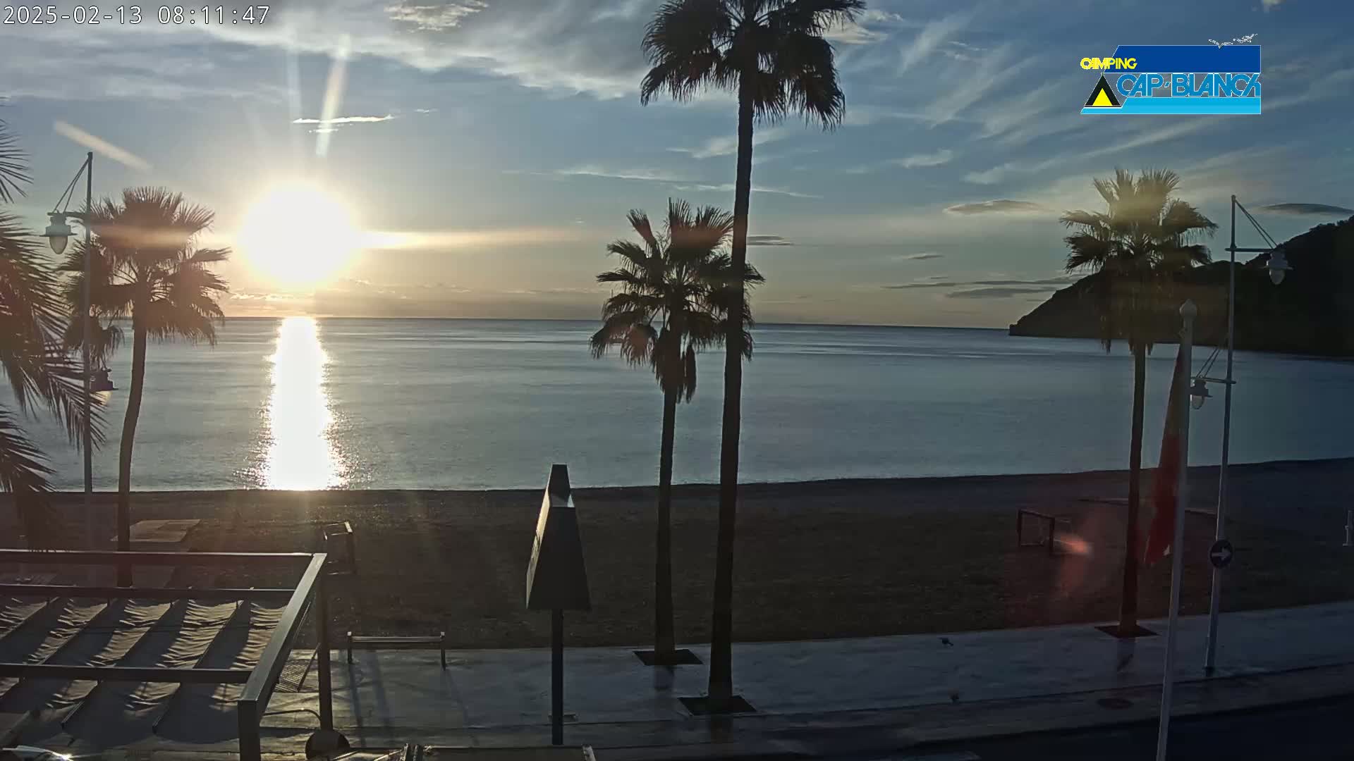 A sunrise over a calm sea, illuminating a sandy beach lined with palm trees and streetlights.