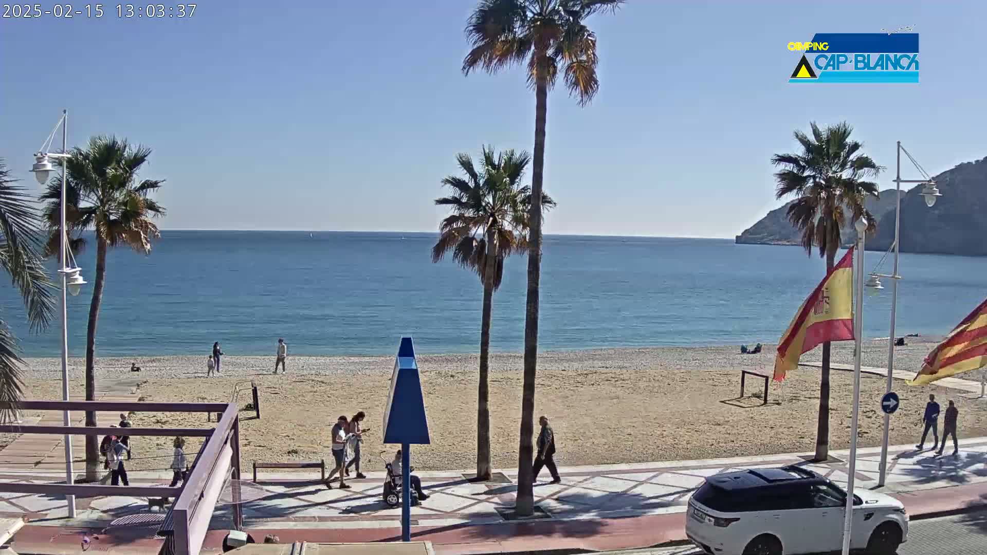 A sunny beach scene shows a calm ocean, sandy beach, palm trees, and people walking along a paved walkway.