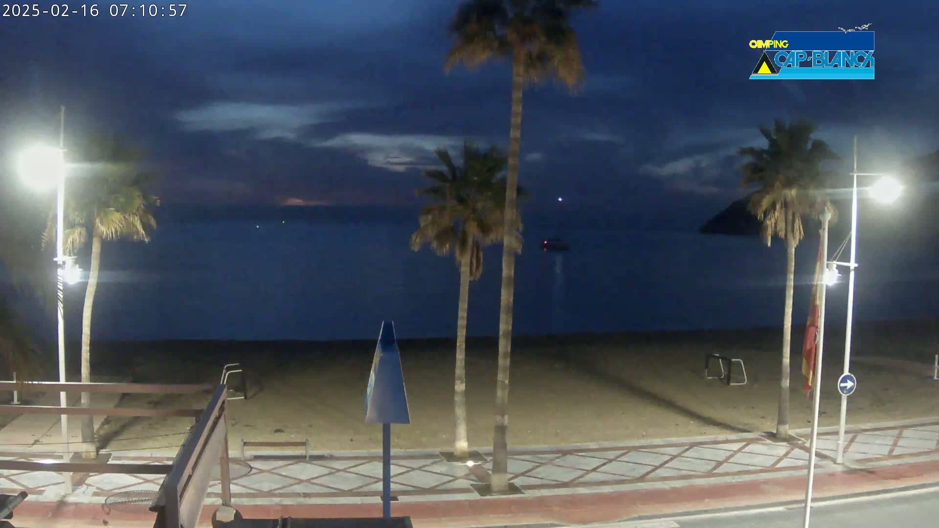 A sandy beach at dusk, under a mostly clear, dark blue sky, is bordered by a paved walkway lined with palm trees and streetlights.