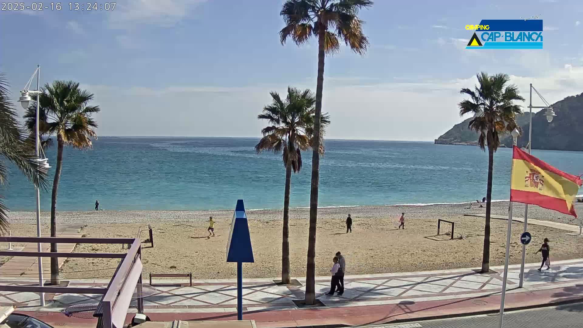 A sandy beach with palm trees, a few people, and a soccer goal is visible next to a calm, blue ocean under a mostly sunny sky.
