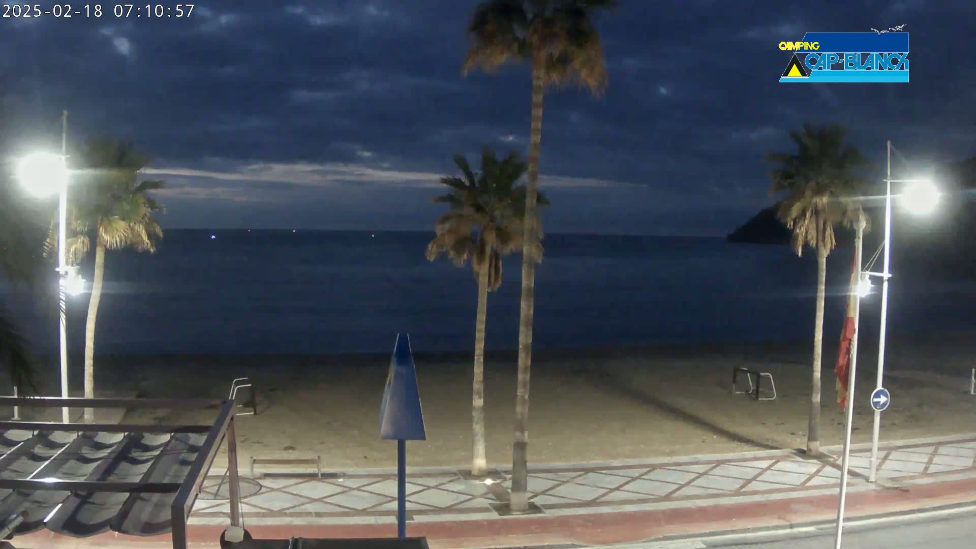 A sandy beach at dusk, under a partly cloudy sky, is lined with palm trees and streetlights, overlooking a calm, dark-blue sea.