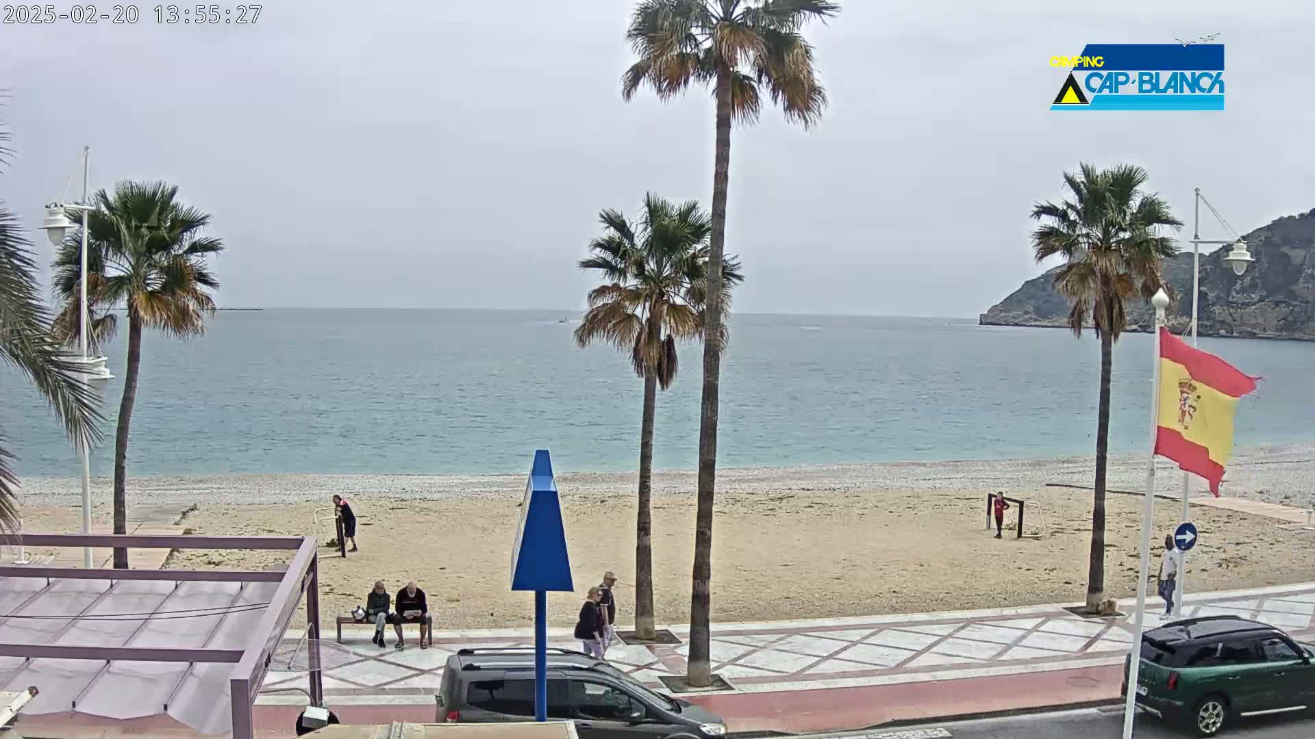 A mostly sunny day at a beach with calm, light-blue water, palm trees, and several people walking along a paved walkway next to a sandy beach.