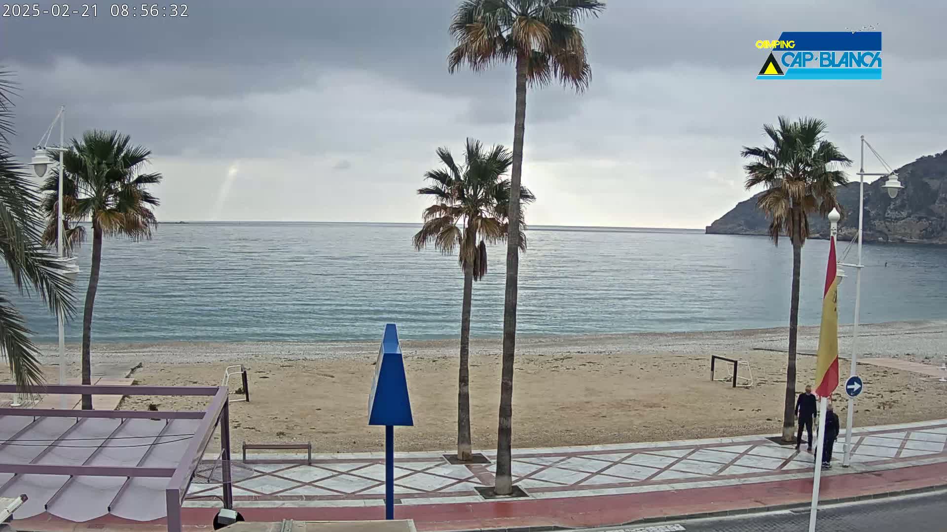 A mostly cloudy day shows a sandy beach with palm trees, a promenade, and two people walking along the shore near a calm sea with a mountain in the background.