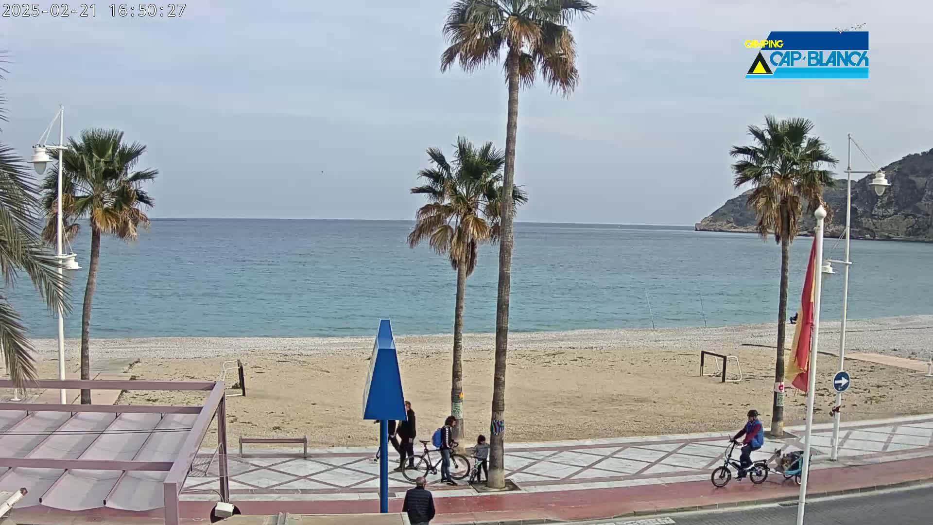A sandy beach with calm blue water is bordered by a paved walkway with several people and bicycles, under a mostly clear sky.