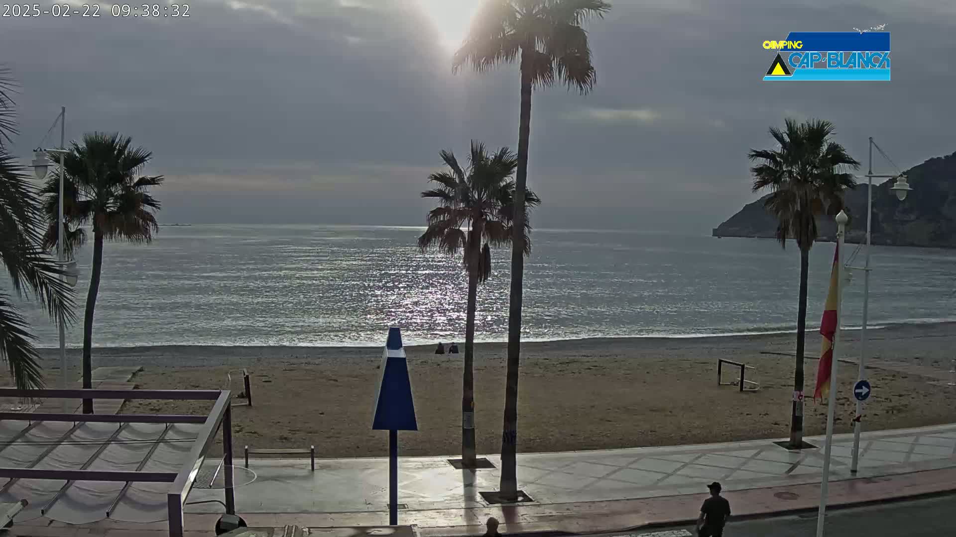 A mostly sunny day shows a sandy beach with palm trees, a calm ocean, and a paved walkway next to the beach.