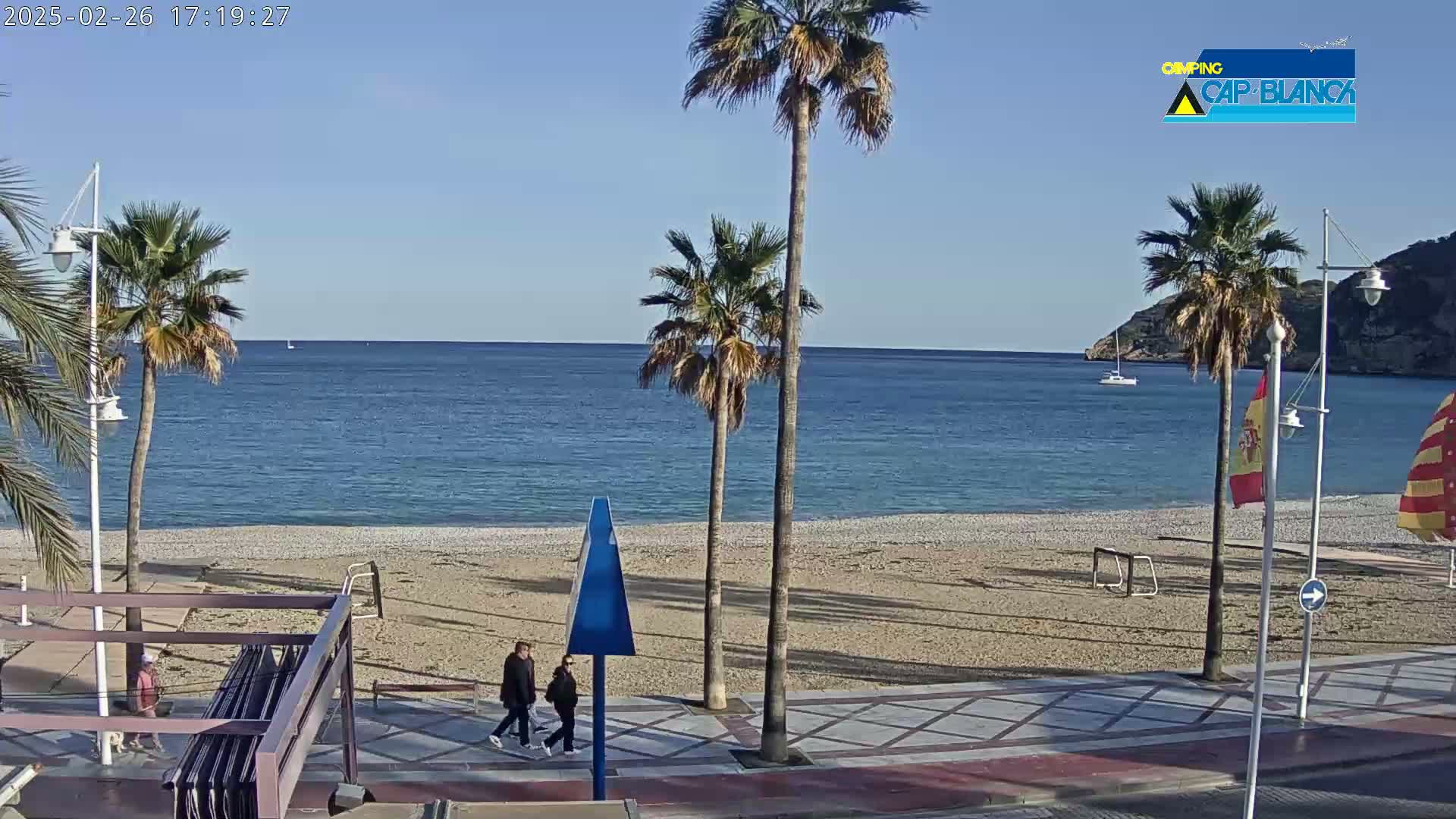 A sunny beach scene shows a calm blue sea, a sandy beach with a few people walking, palm trees lining a paved walkway, and a small boat in the distance.