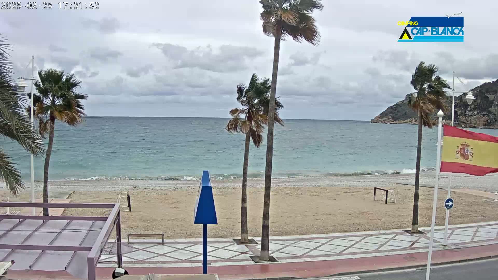 A sandy beach with palm trees is seen on an overcast day, next to a calm, blue-green sea and a rocky cliff in the distance.
