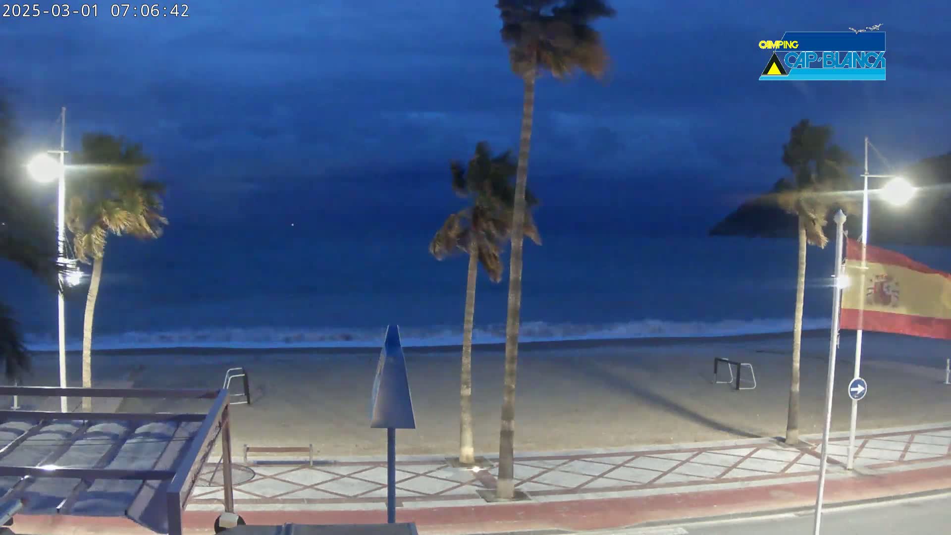 A windy, twilight scene shows a sandy beach with palm trees, streetlights, and a Spanish flag, overlooking a dark blue ocean with small waves.