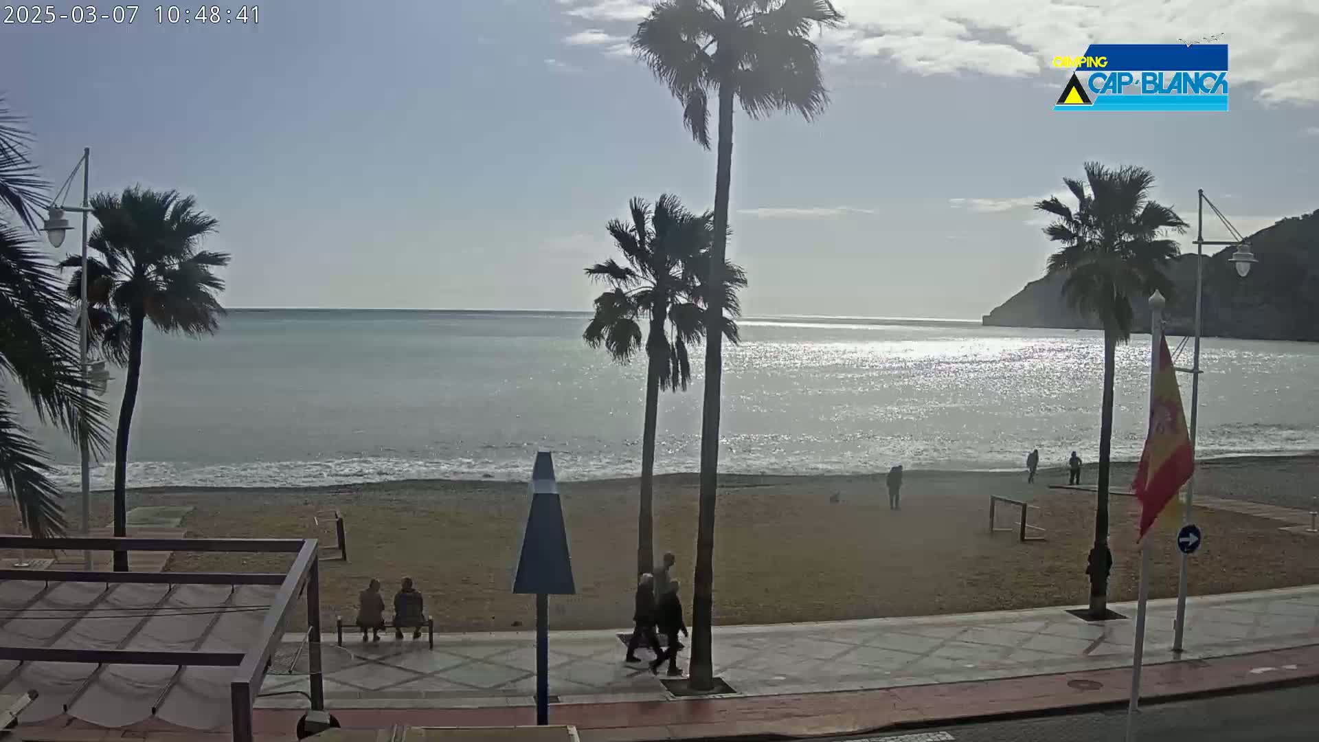 A mostly sunny day at a beach with palm trees, a sandy shore, calm waves, and several people walking along a paved walkway.