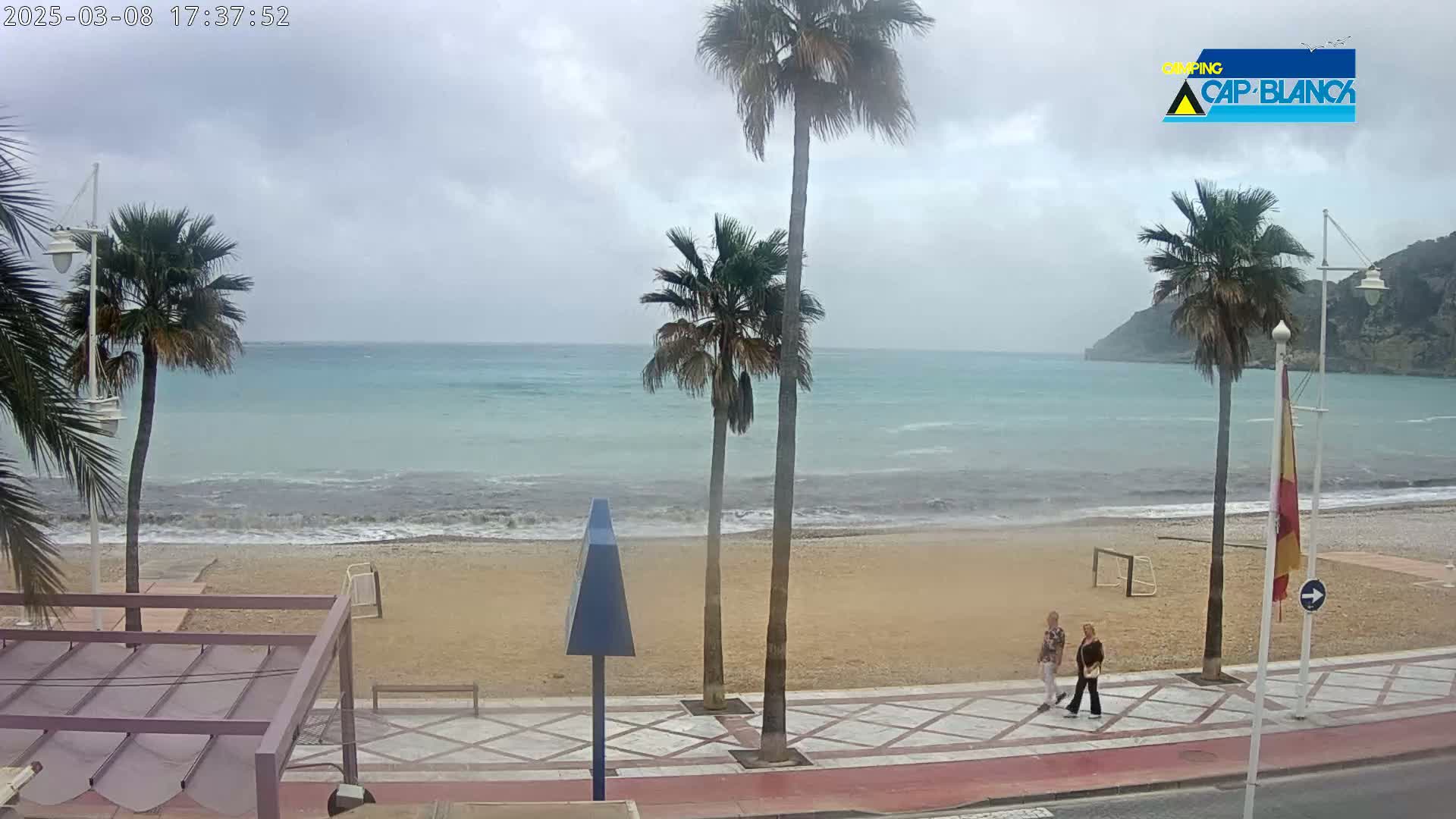 A sandy beach with palm trees and two people walking along a paved walkway, overlooks a somewhat choppy turquoise ocean under an overcast sky.