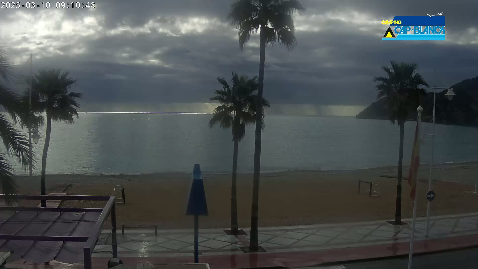 A mostly cloudy day reveals a calm sea beside a sandy beach, palm trees, and a paved walkway.