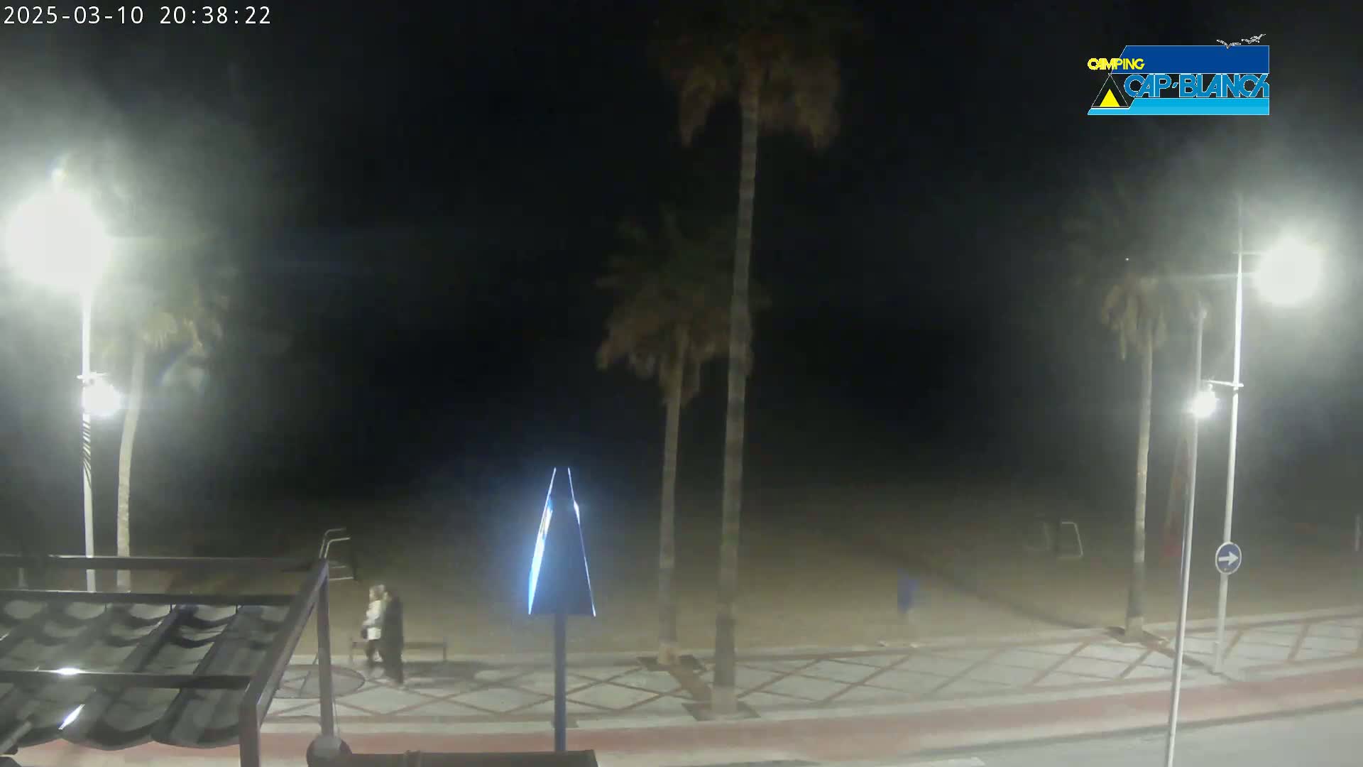 A nighttime beach scene shows a hazy, dimly lit sandy beach with palm trees, streetlights, and a few people walking along a paved walkway.