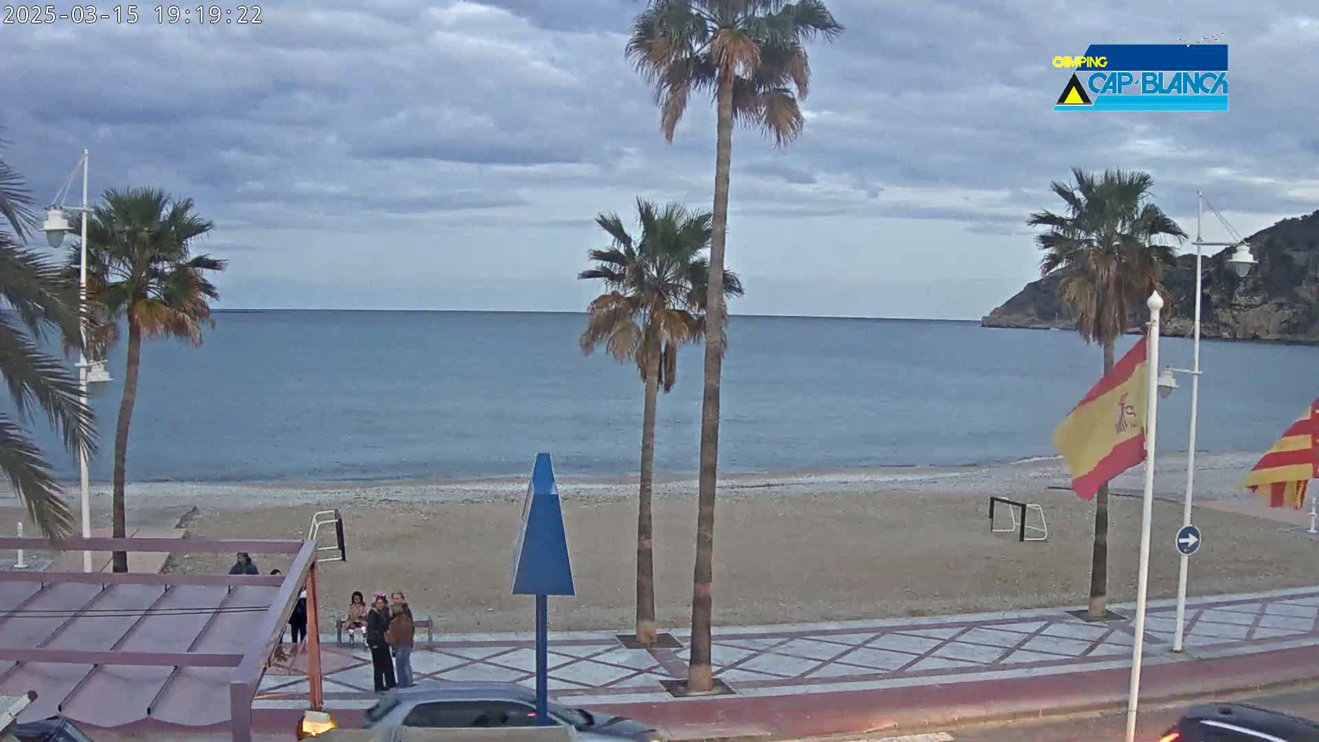 A mostly cloudy day shows a beach scene with palm trees, a calm sea, and several people standing near a walkway.