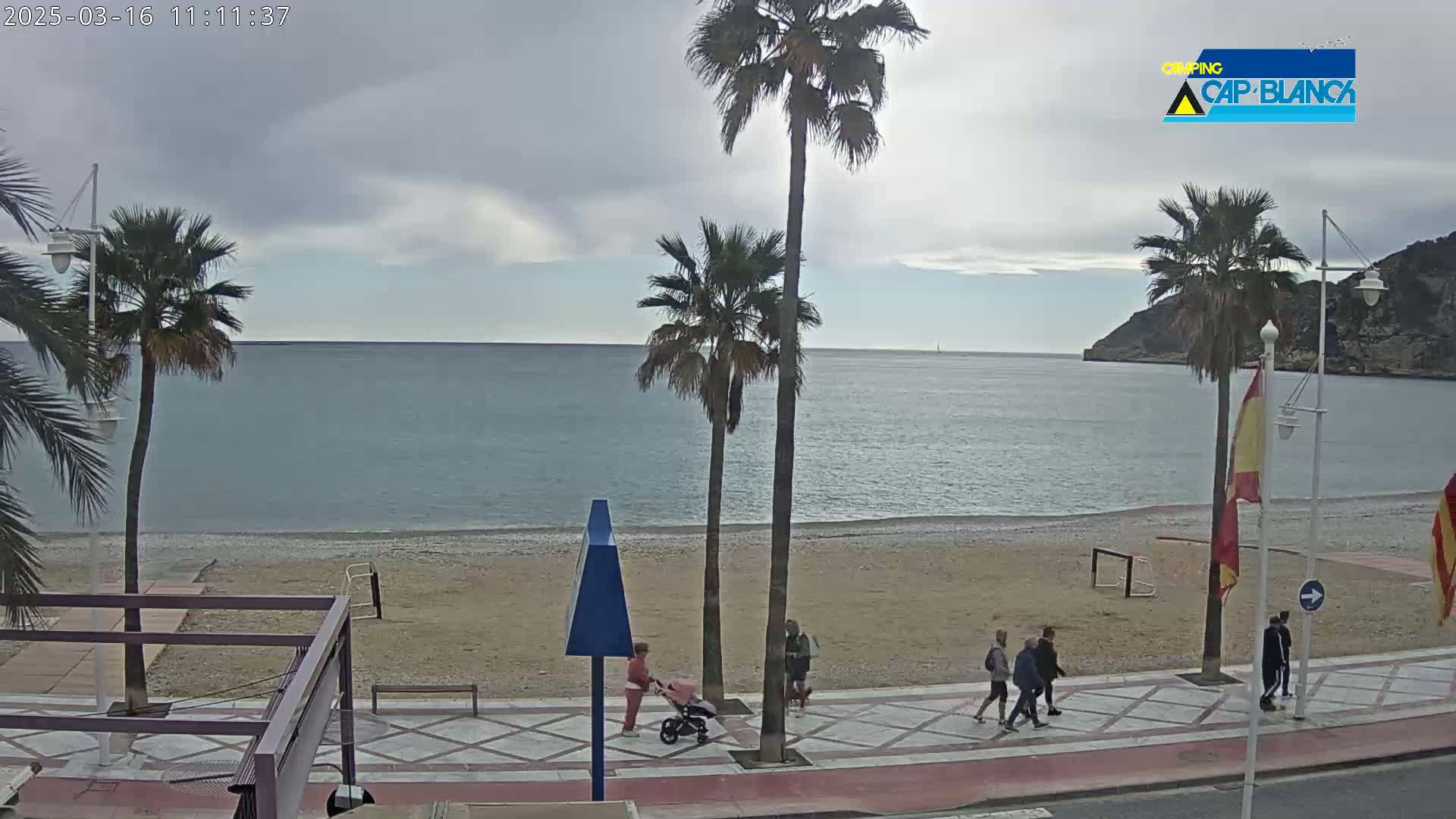 A mostly cloudy day shows a sandy beach with calm ocean water, palm trees, and a few people walking along a paved walkway.