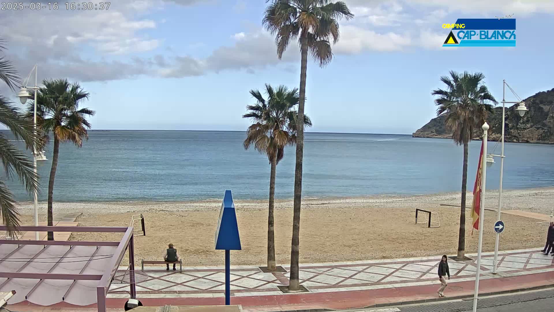 A mostly sunny day at a beach with palm trees, a sandy shore, calm water, and a few people walking along the boardwalk.