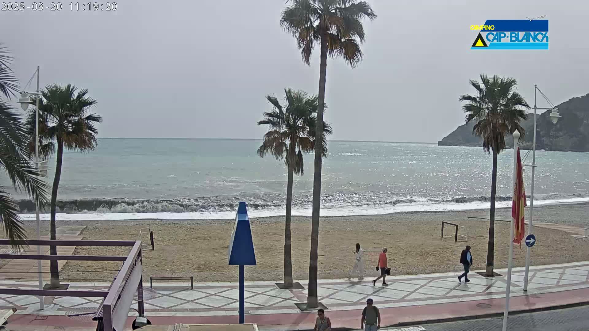 A sandy beach with moderate waves under an overcast sky is viewed from a promenade, with several palm trees and a few people walking along the walkway.