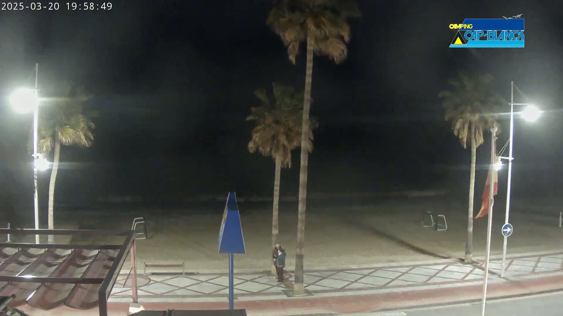 A nighttime beach scene shows a sandy beach, palm trees, and two people standing on a paved walkway under bright streetlights.
