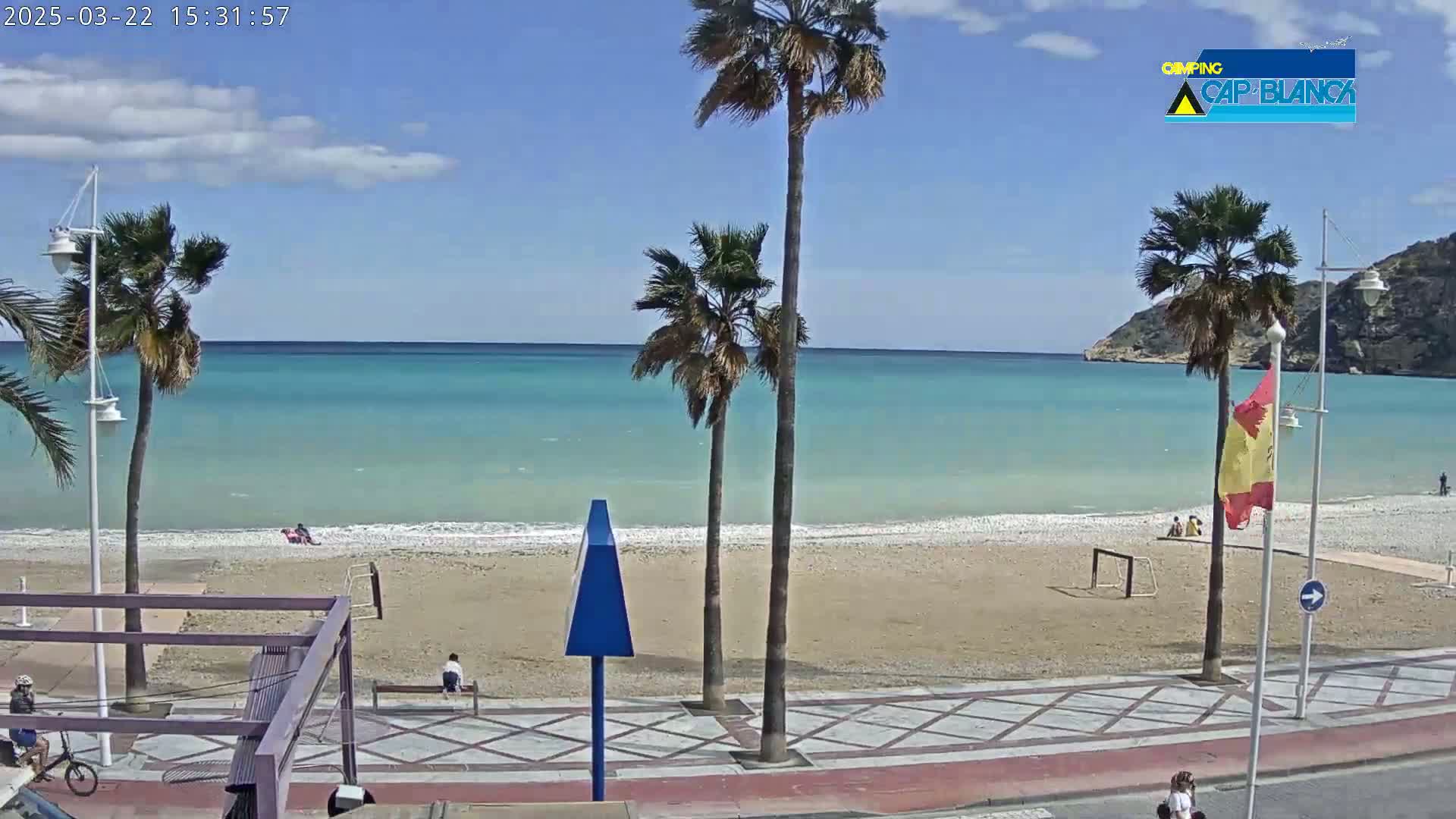 A sandy beach with calm, turquoise water is viewed from a promenade, under a mostly sunny sky with a few clouds.