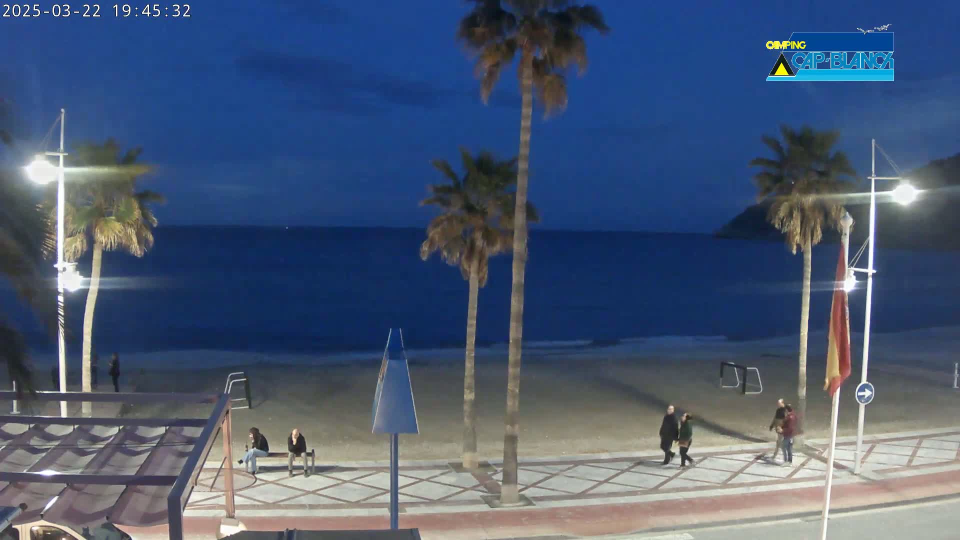 A sandy beach at dusk, under a dark blue twilight sky, is shown with several palm trees, a few people walking along the boardwalk, and a calm ocean.