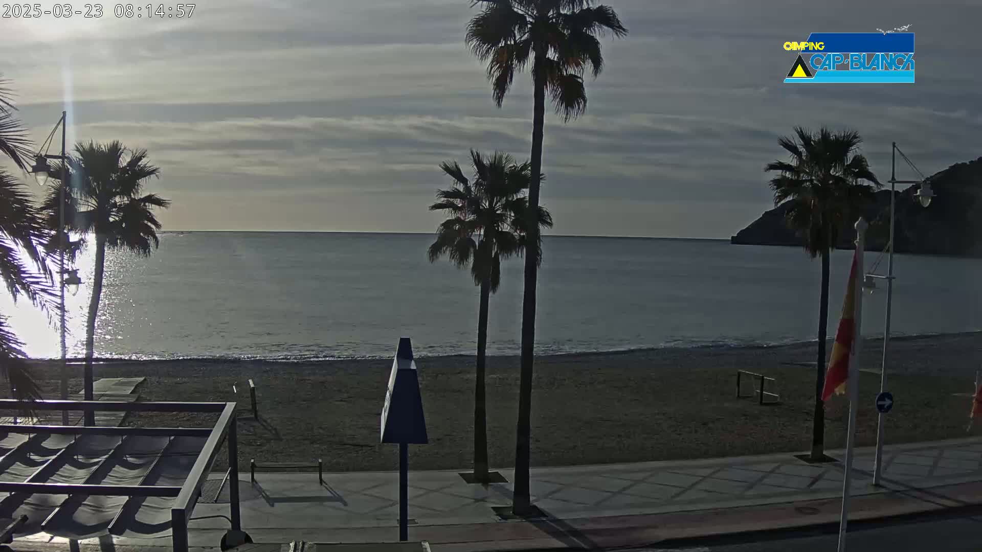 A mostly cloudy sunrise illuminates a calm ocean beach with palm trees and a paved walkway.