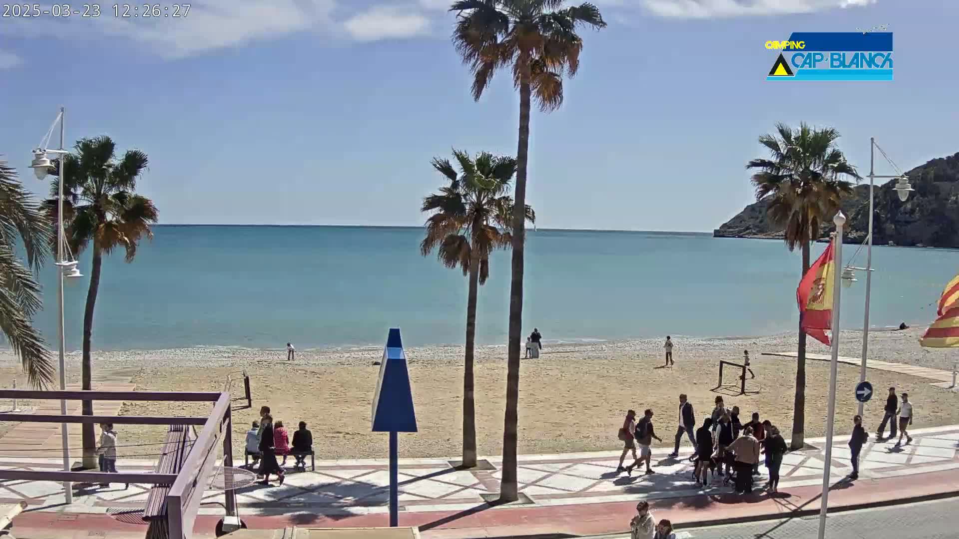 A sandy beach with calm, light-blue water is bordered by a paved walkway with people walking and sitting, under a sunny sky and palm trees.