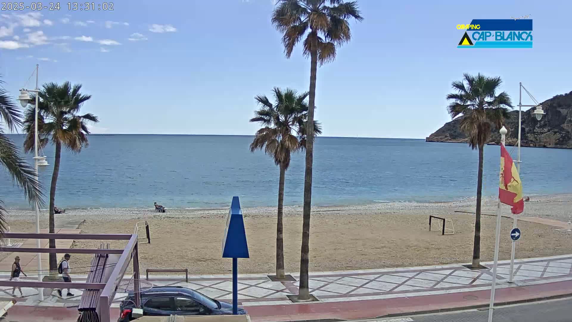 A sandy beach with calm blue water is bordered by a paved walkway, palm trees, and a few people; the sky is mostly clear and sunny.