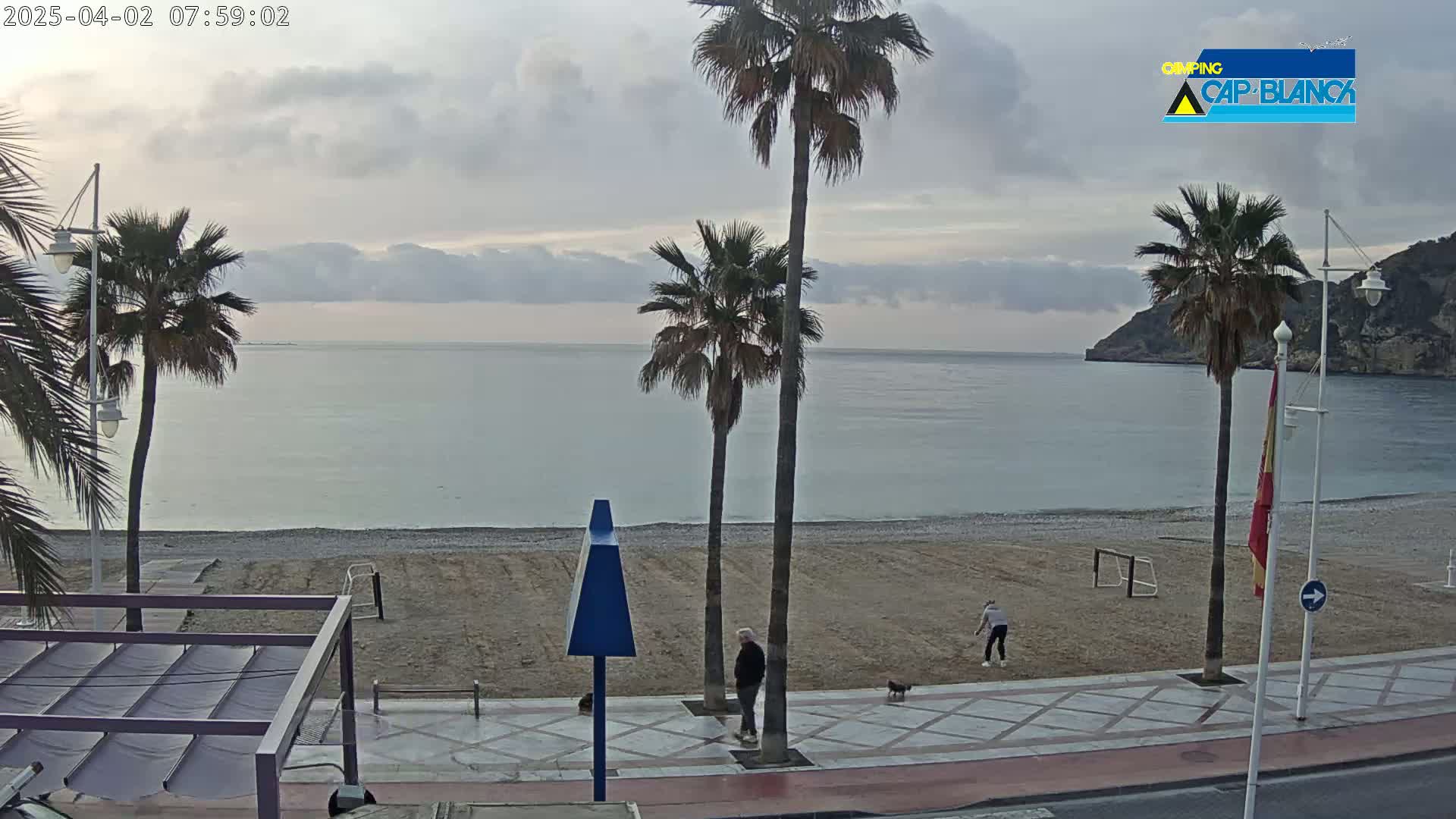 A mostly cloudy day overlooks a calm, light-blue sea with a sandy beach, palm trees, and a few people walking along a paved walkway.