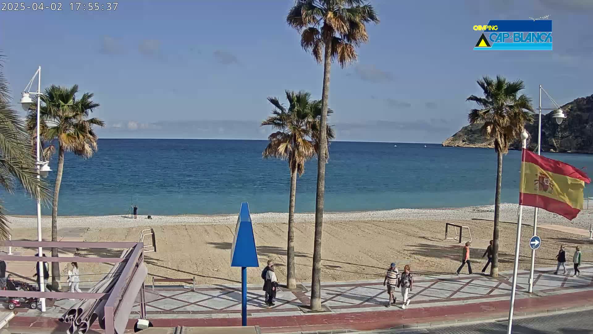 A mostly sunny day at a beach with palm trees, a few people walking on the boardwalk and a sandy beach.
