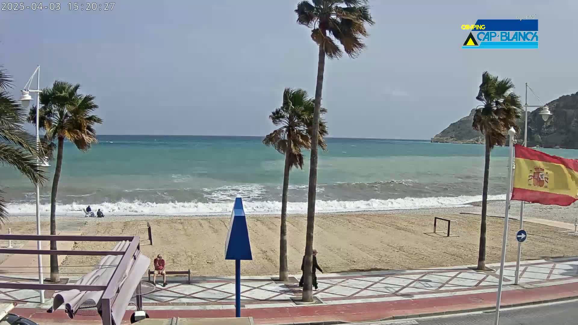 A sandy beach with palm trees and a few people, bordered by a paved walkway, under a mostly clear sky with some wind causing waves in the ocean.