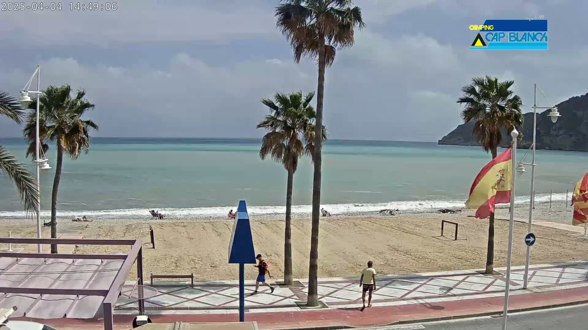 A mostly sunny beach scene shows a sandy beach with a few people, palm trees, and a calm sea with small waves under a partly cloudy sky.