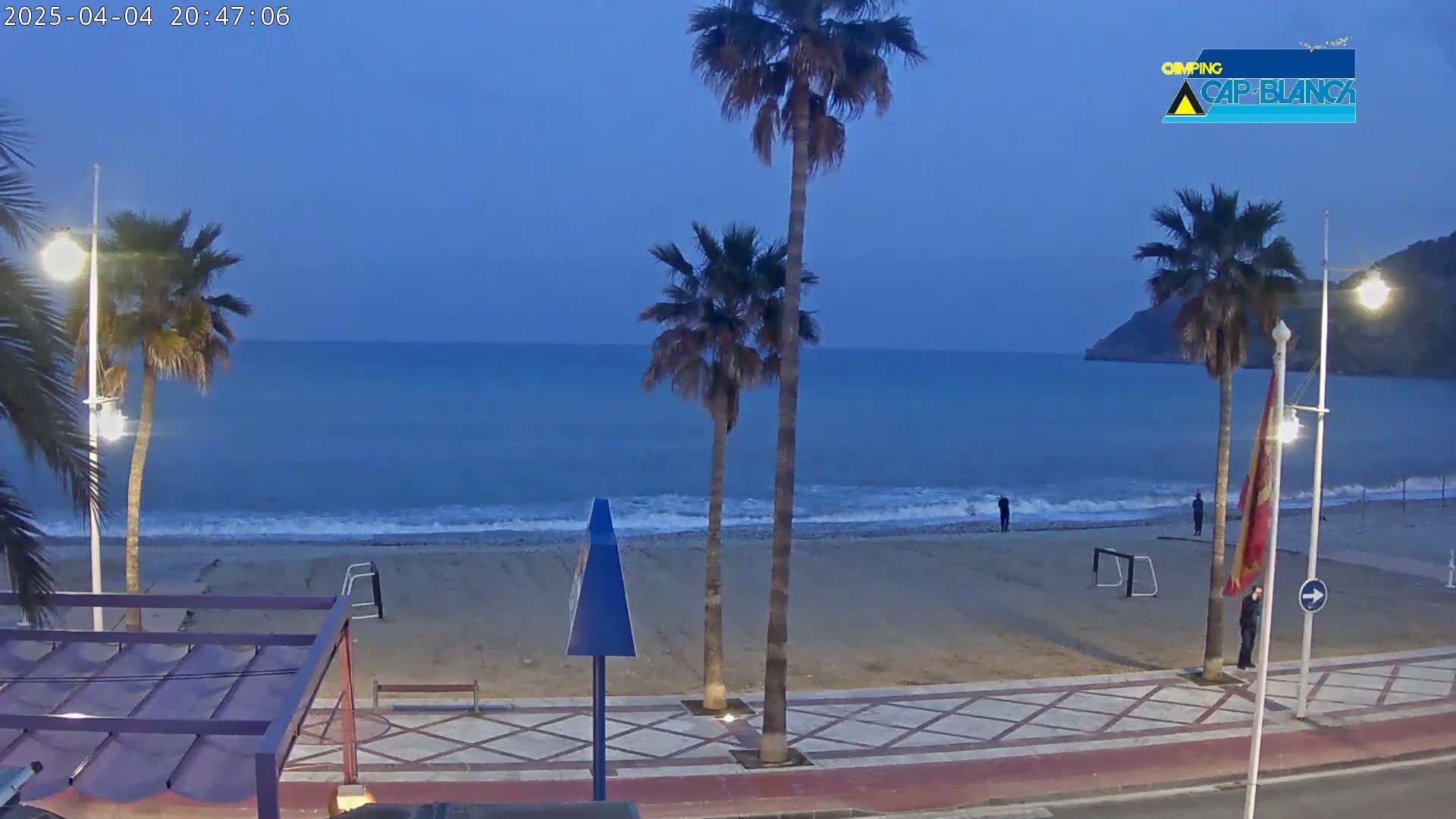 A sandy beach at twilight is bordered by palm trees and a paved walkway, with a calm sea in the background.