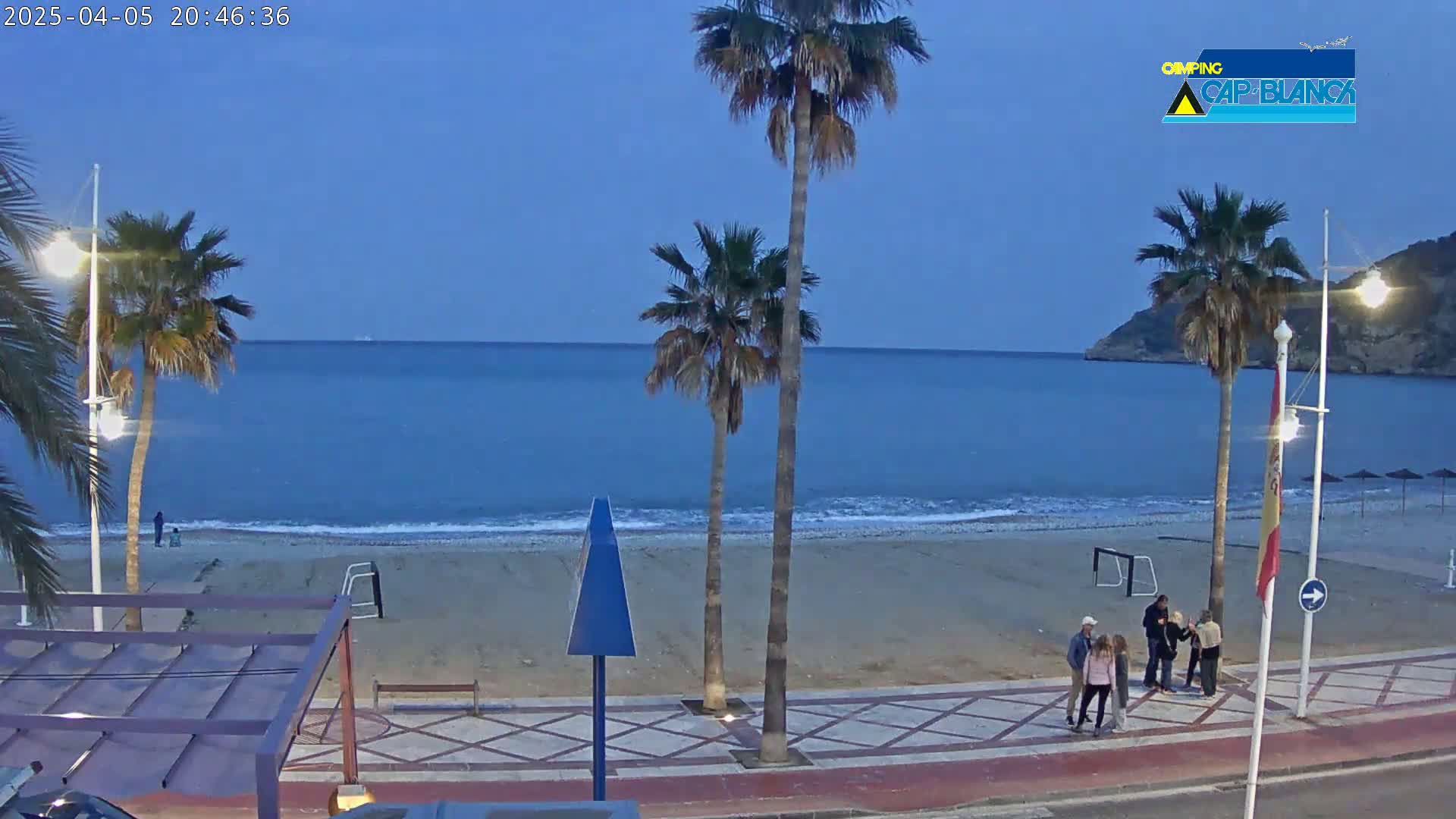 A mostly empty beach at dusk, with palm trees and a few people gathered on the boardwalk next to the calm ocean.