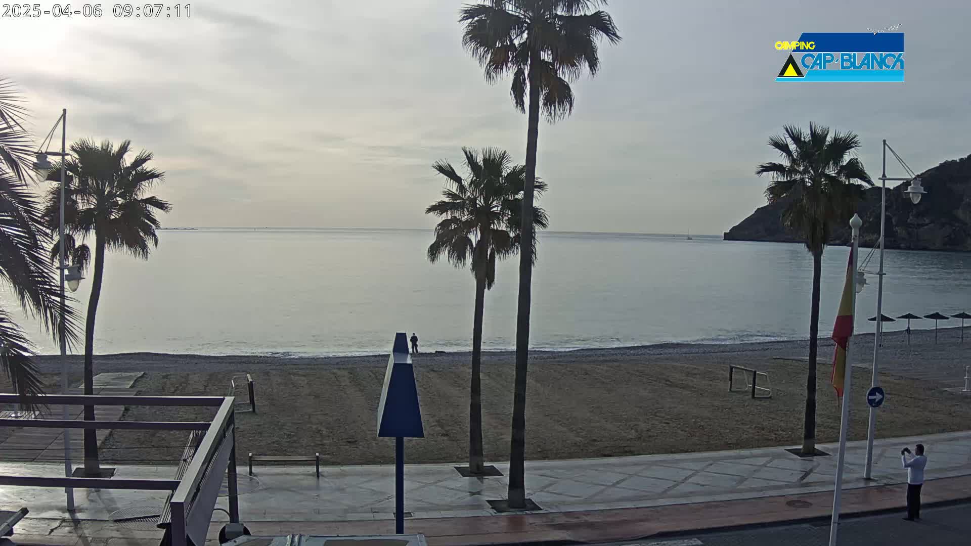A mostly cloudy day shows a beach with palm trees, a calm sea, and a person taking a picture.