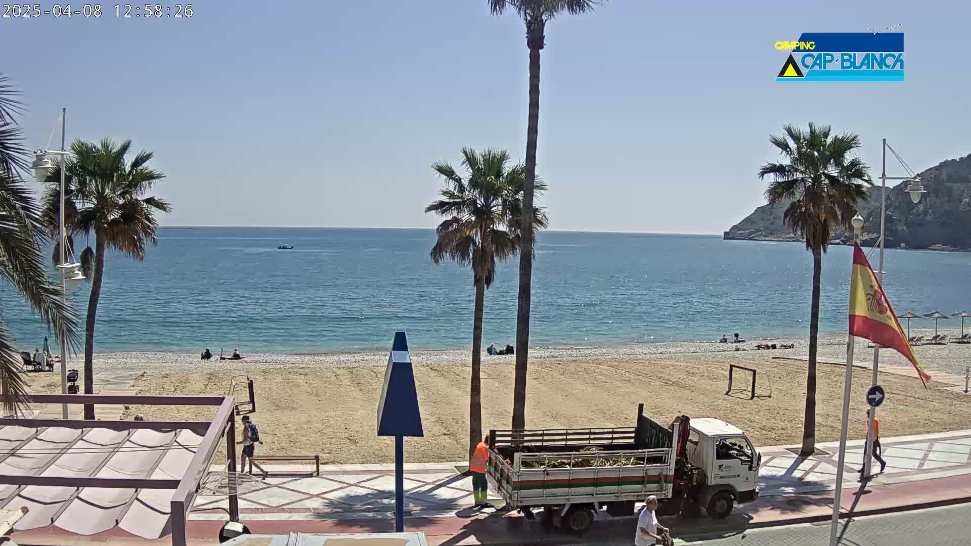 A sunny beach scene shows a partly populated sandy beach next to a calm blue sea, with palm trees and a pickup truck on the promenade.