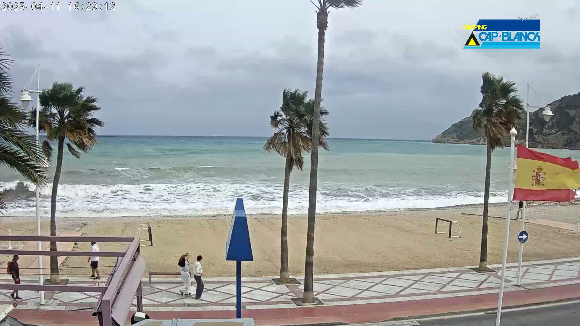 A windy, overcast day at a beach with palm trees, a few people walking, and moderate waves breaking on the shore.