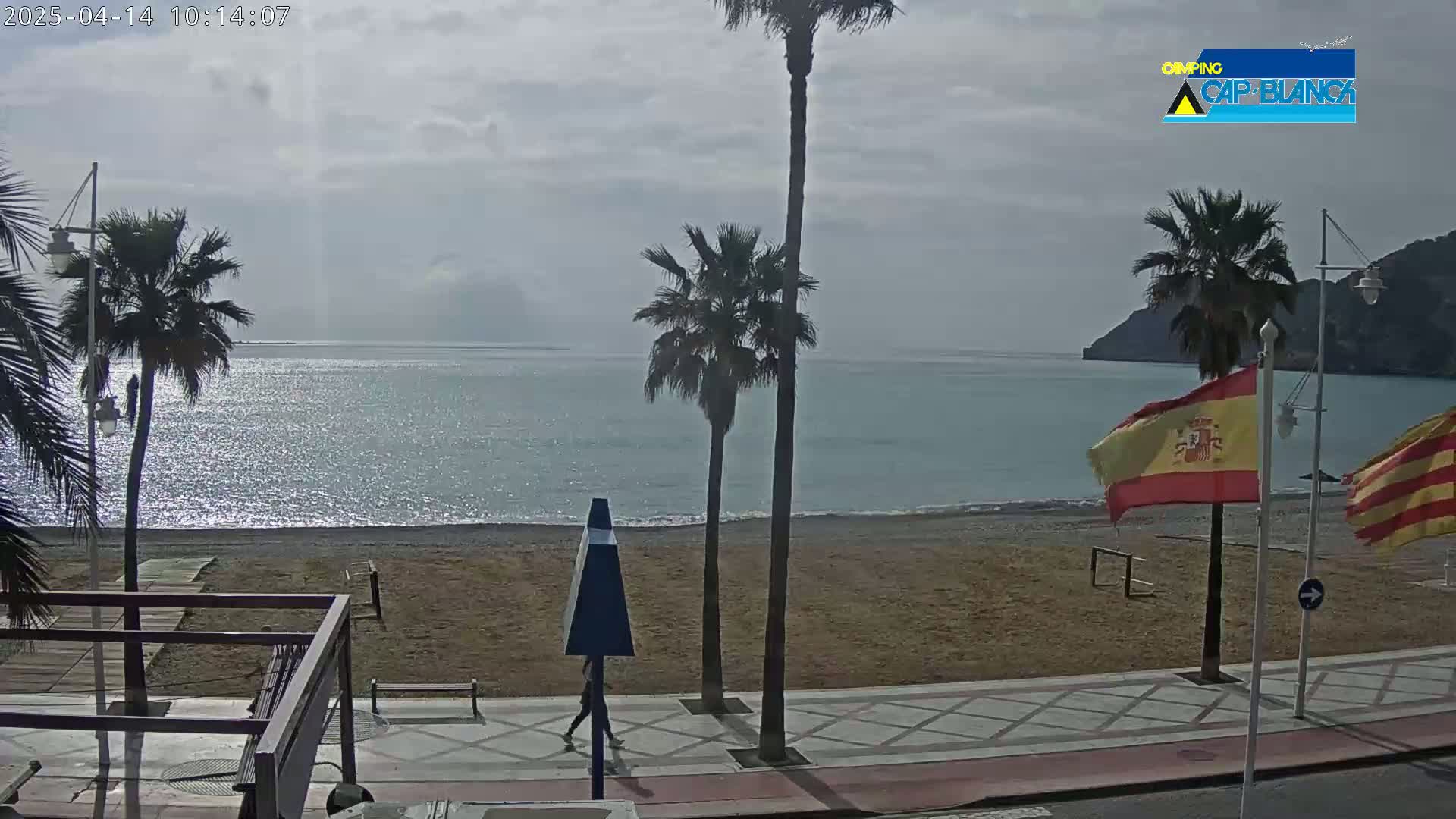A mostly sunny day reveals a sandy beach, calm ocean, palm trees, and Spanish flags along a paved walkway.