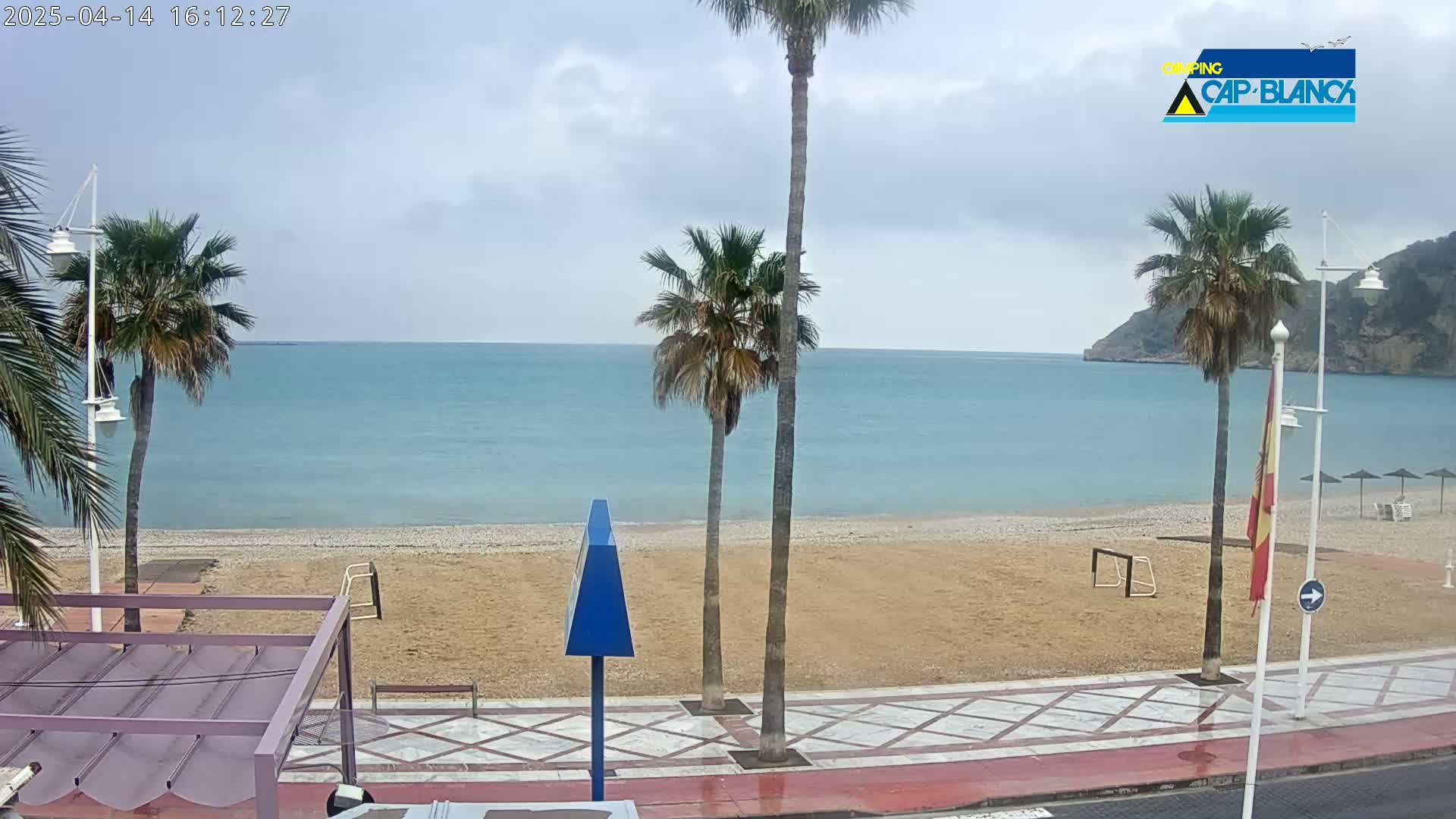 A sandy beach with palm trees and a calm, light-blue sea under an overcast sky is visible from a seaside promenade.