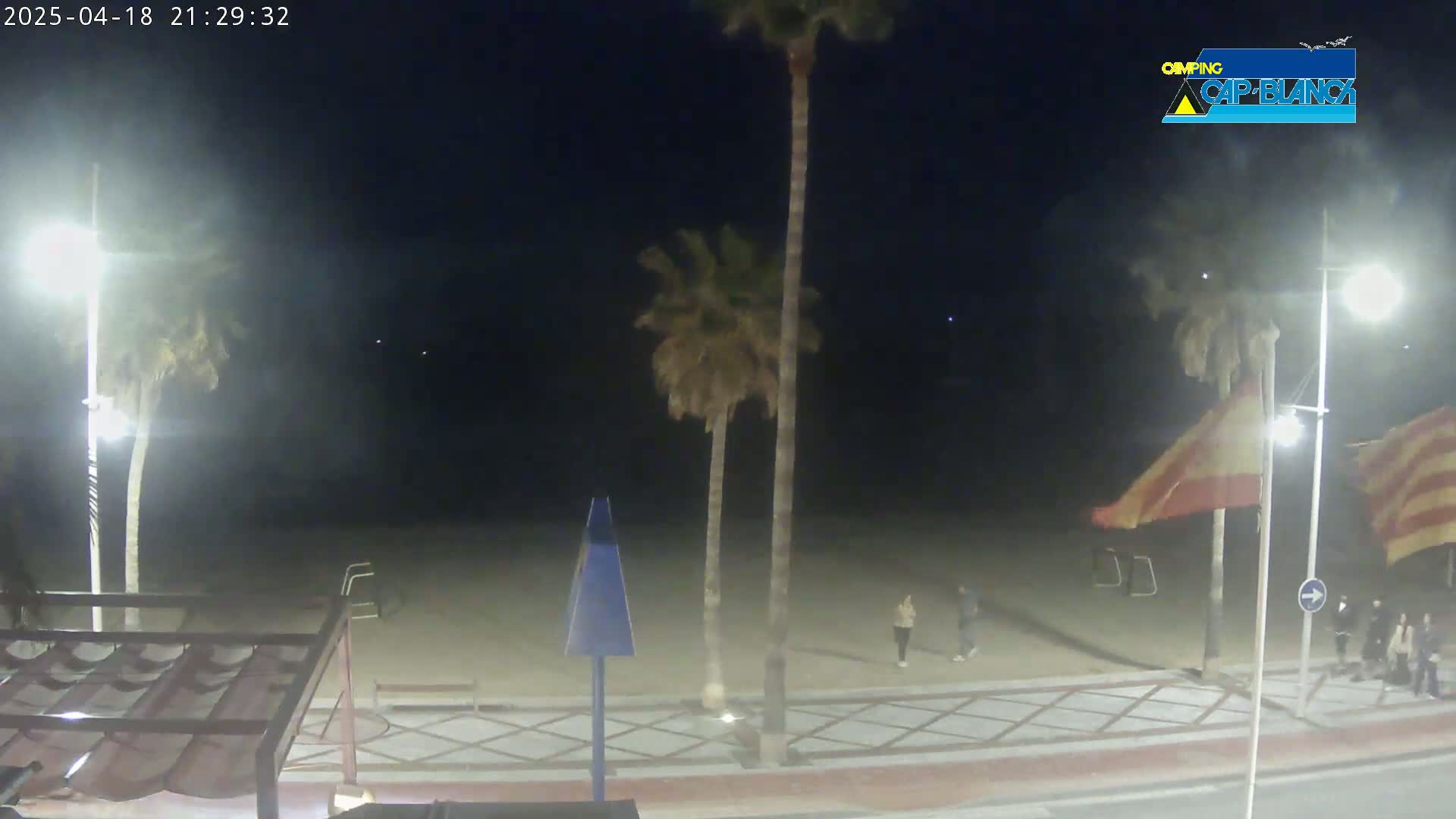 At night, a mostly empty beach is lit by streetlights, with a few people walking near palm trees and flags.