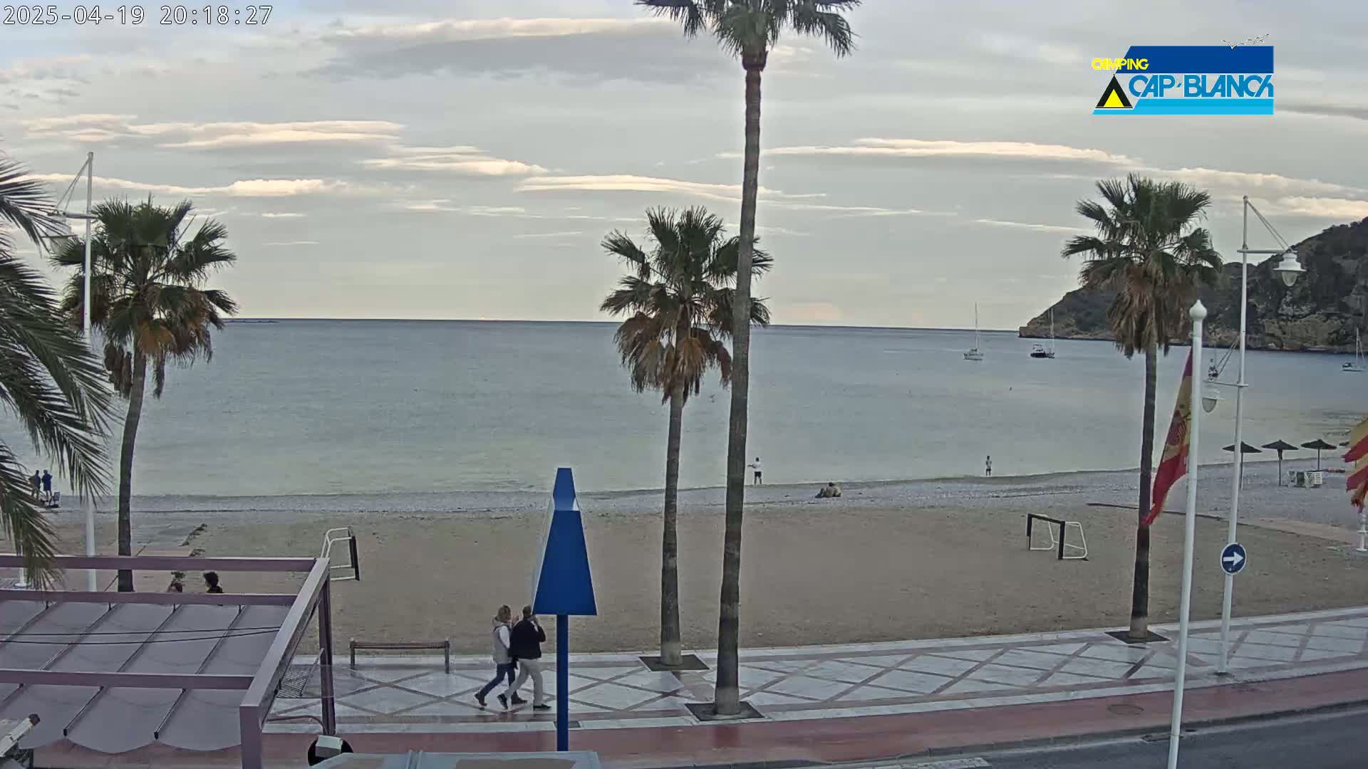 A mostly cloudy evening scene shows a sandy beach with palm trees, a calm sea, and a few people walking along a promenade.