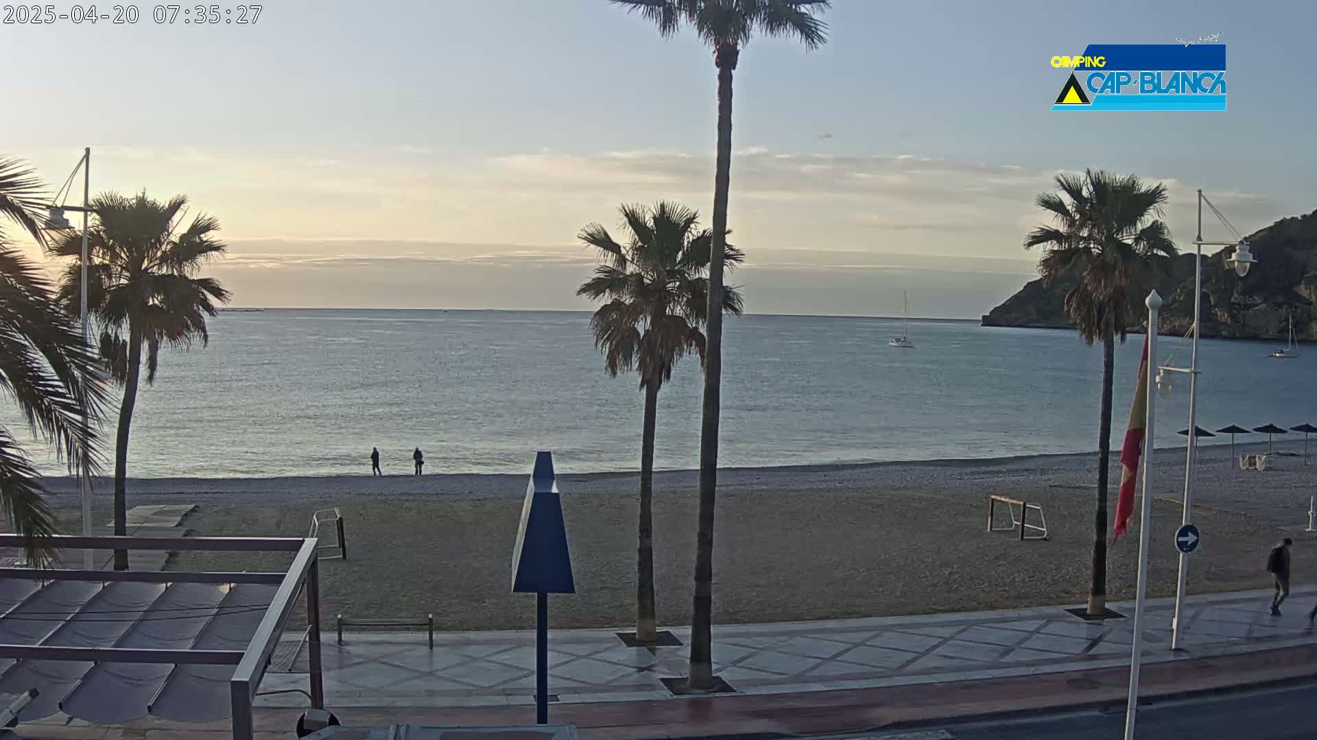 A calm, sunrise-lit ocean beach with several palm trees and a few people walking along the shoreline.