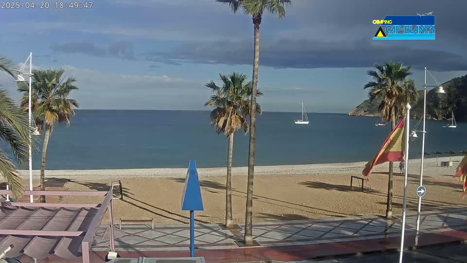 A mostly sunny day at a beach with palm trees, a sandy shore, calm blue water, and a few sailboats in the distance.