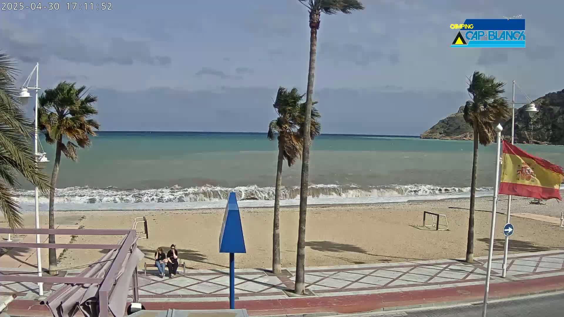 A windy day at a beach shows palm trees bending in the breeze, waves crashing on the shore, and two people sitting on a bench near a sandy beach and boardwalk.